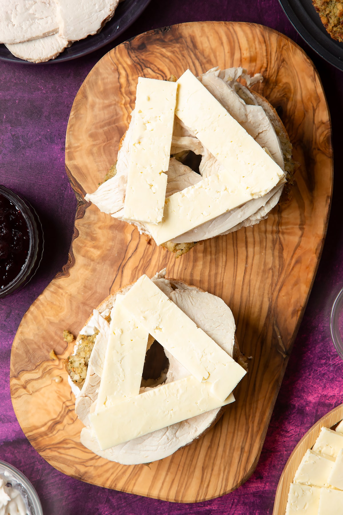 Overhead shot of halved bagel with spread on topped with stuffing slices, turkey and cheese on a wooden plate
