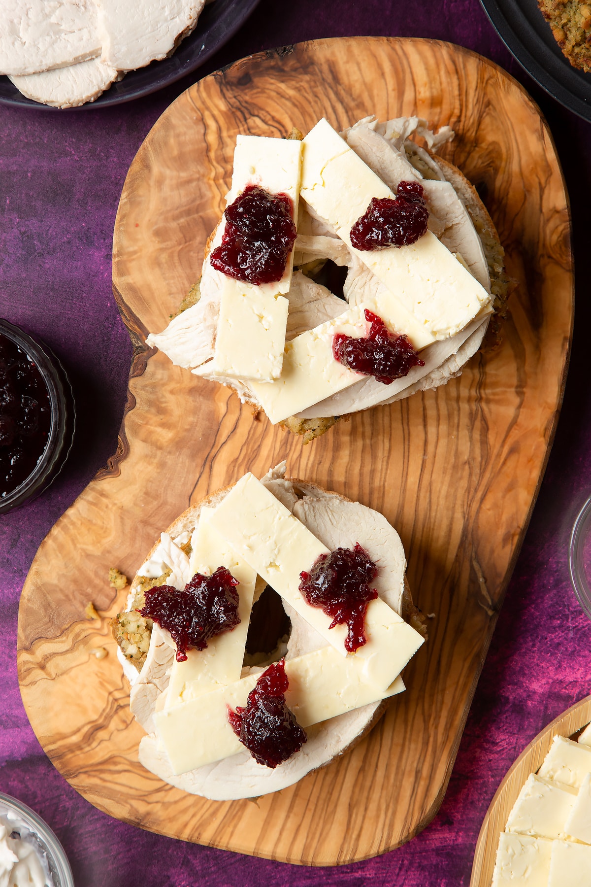 Overhead shot of halved bagel with spread on topped with stuffing slices, turkey, cheese and cranberry sauce on a wooden plate