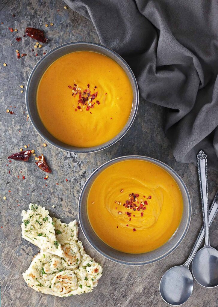A grey bowls filled with Roasted Sweet Potato & Ginger Soup on a dark grey background. The sides are decorated with spoons and chilli flakes.