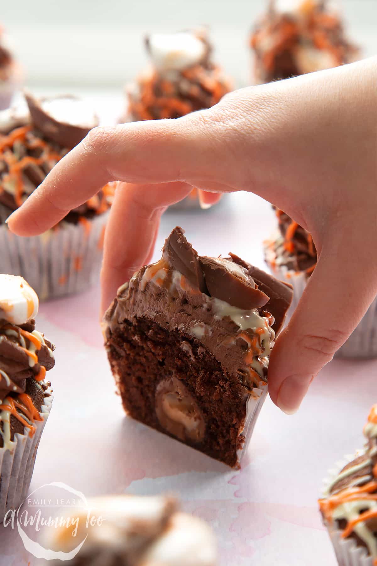 Cadbury Creme Egg cakes on a pink surface. A cake reaches for one that has been cut in half, showing the hidden Creme Egg Mini inside.