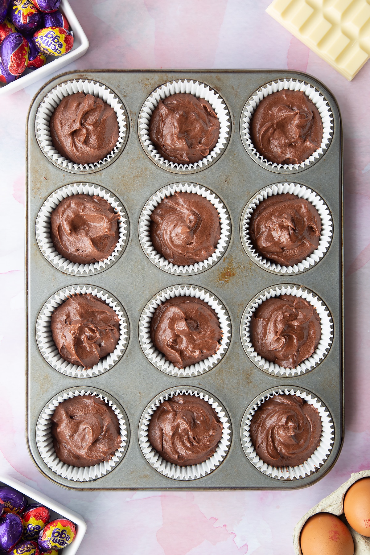 Chocolate cake batter divided into a 12 hole muffin tray lined with cases. Ingredients to make Cadbury Creme Egg cakes surround the tray.