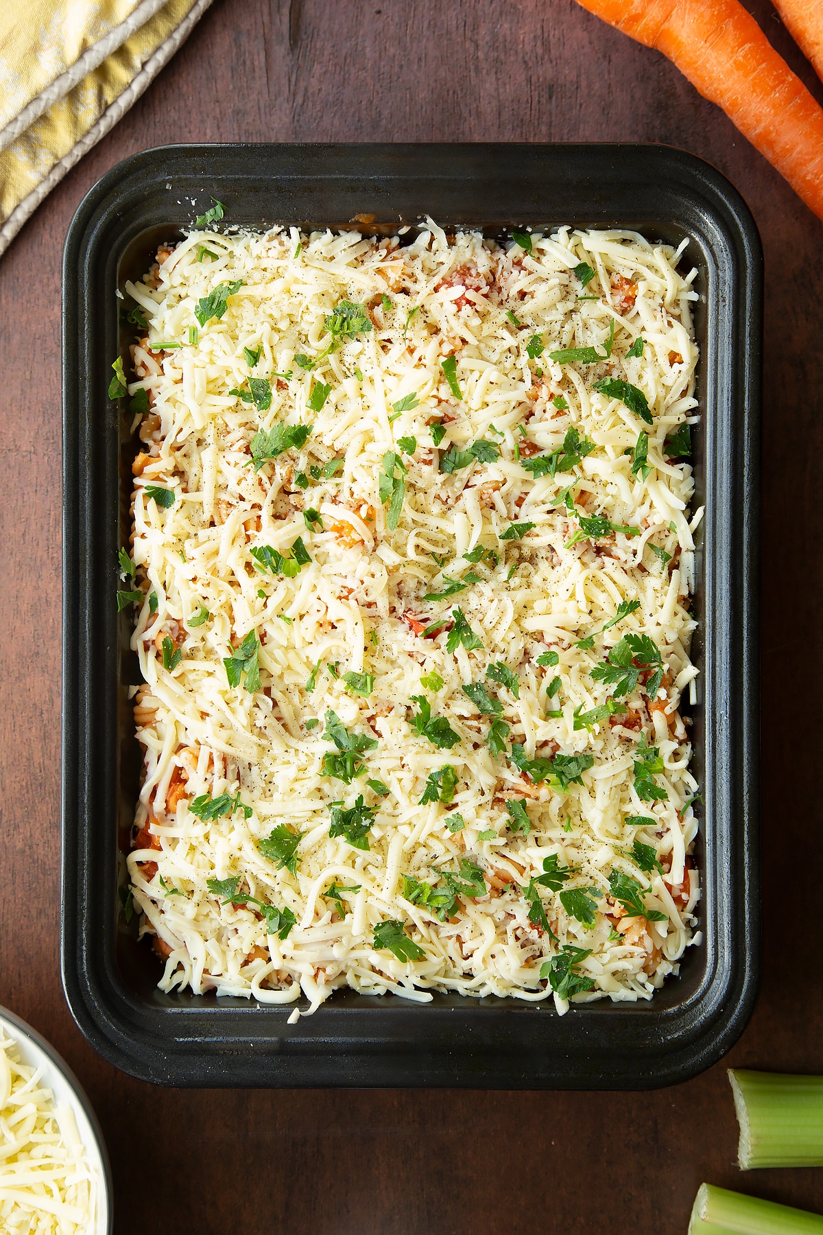 Baked beans and pasta in a tray, topped with shredded mozzarella, parsley and pepper. Ingredients to make pasta and baked beans surround the tray.
