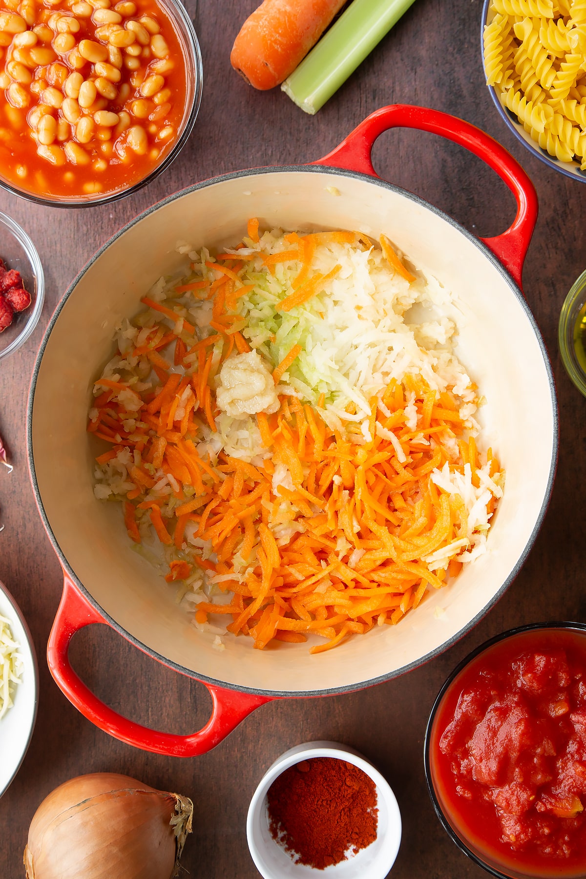 Olive oil, carrot, onion, celery and garlic in a saucepan. Ingredients to make pasta and baked beans surround the pan.