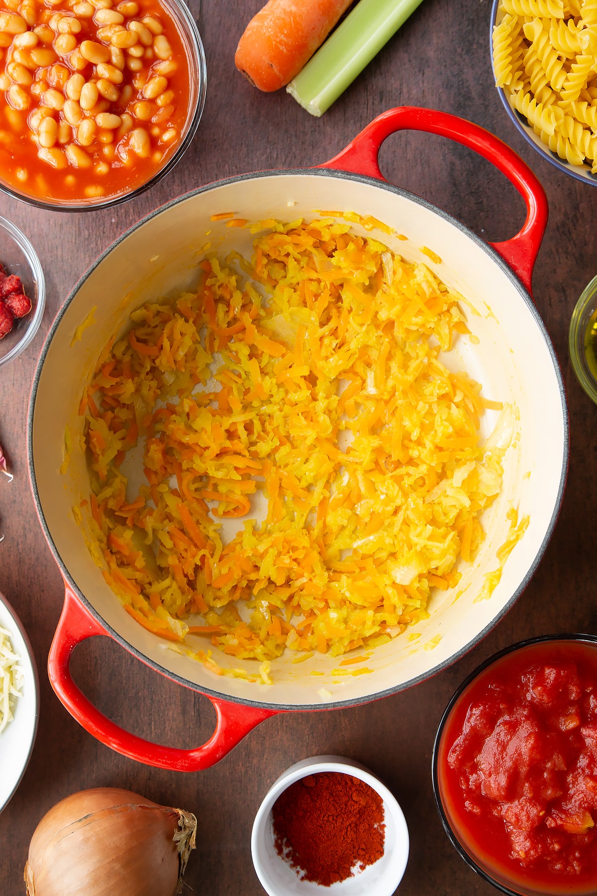 Fried carrot, onion, celery and garlic in a saucepan. Ingredients to make pasta and baked beans surround the pan.