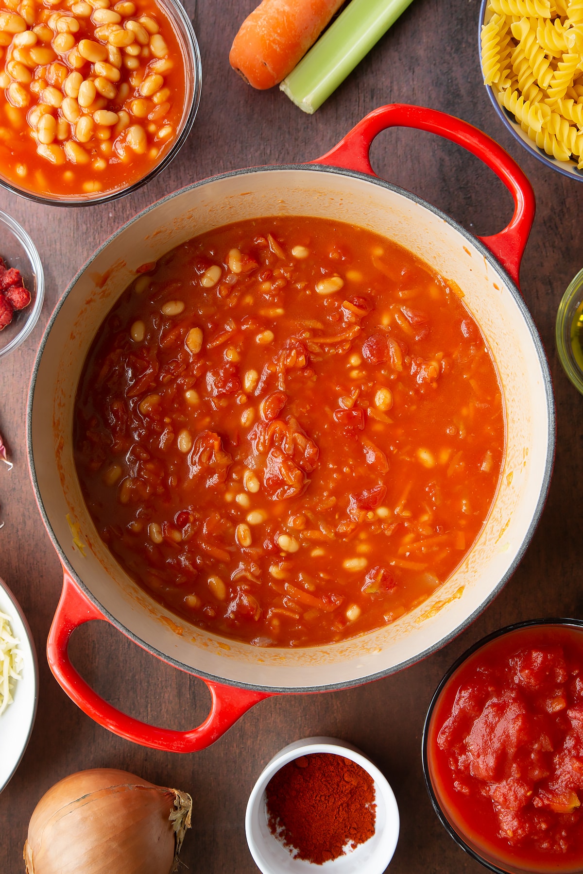 Grated veg, baked beans, tomatoes, tomato puree, paprika and water in a saucepan. Ingredients to make pasta and baked beans surround the pan.