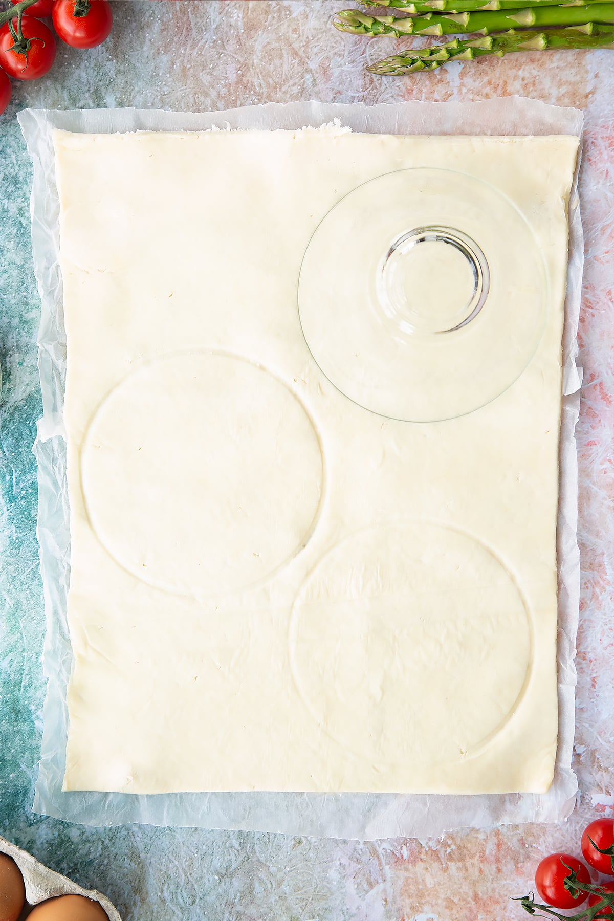 A sheet of shortcrust pastry with an upturned bowl on it. Three circles have been marked. Ingredients to make asparagus tartlets surround the pastry.