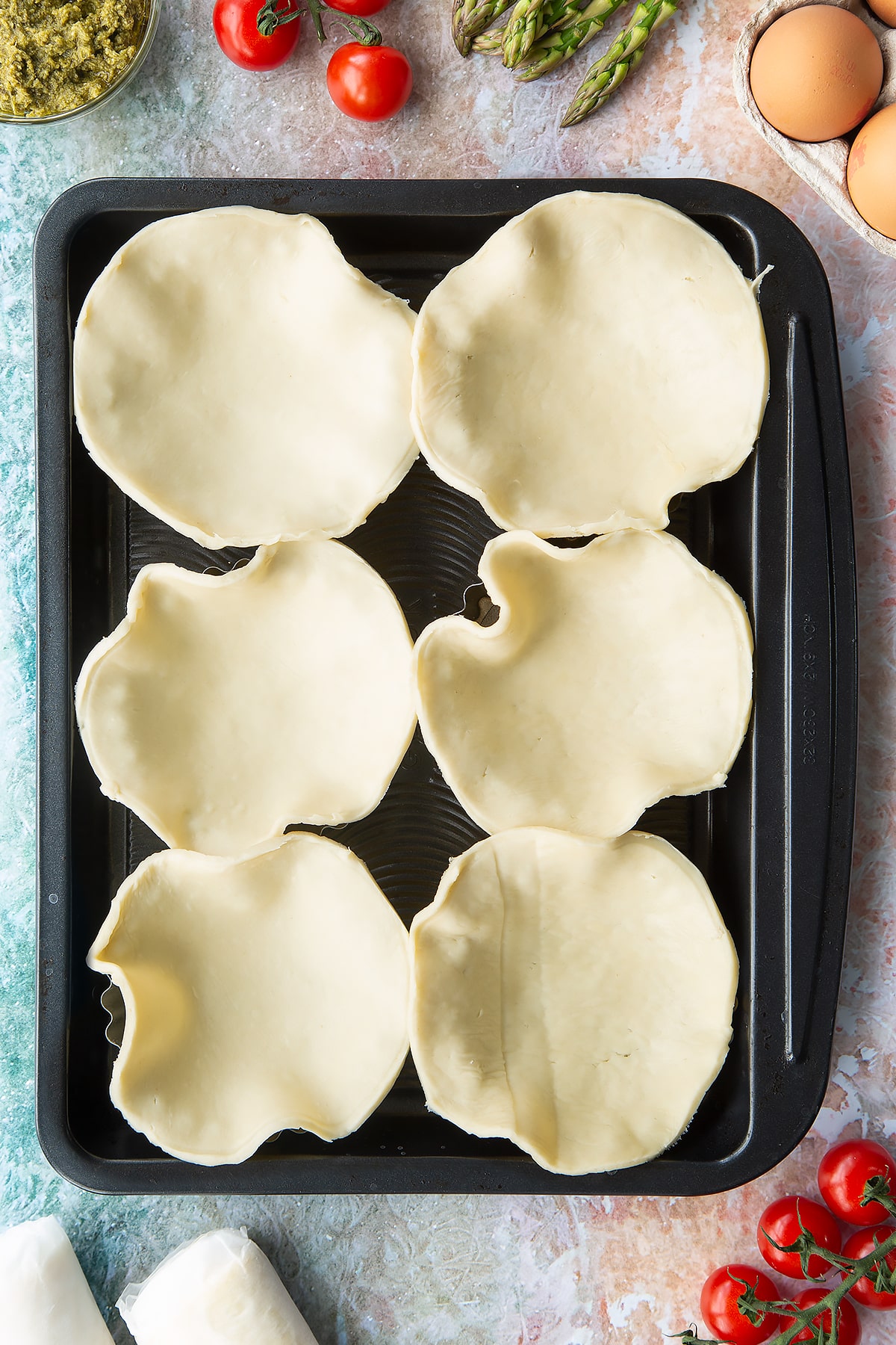 Six tartlet tins on a baking tray. Pastry discs sit loosely in the tins. Ingredients to make asparagus tartlets surround the tray.