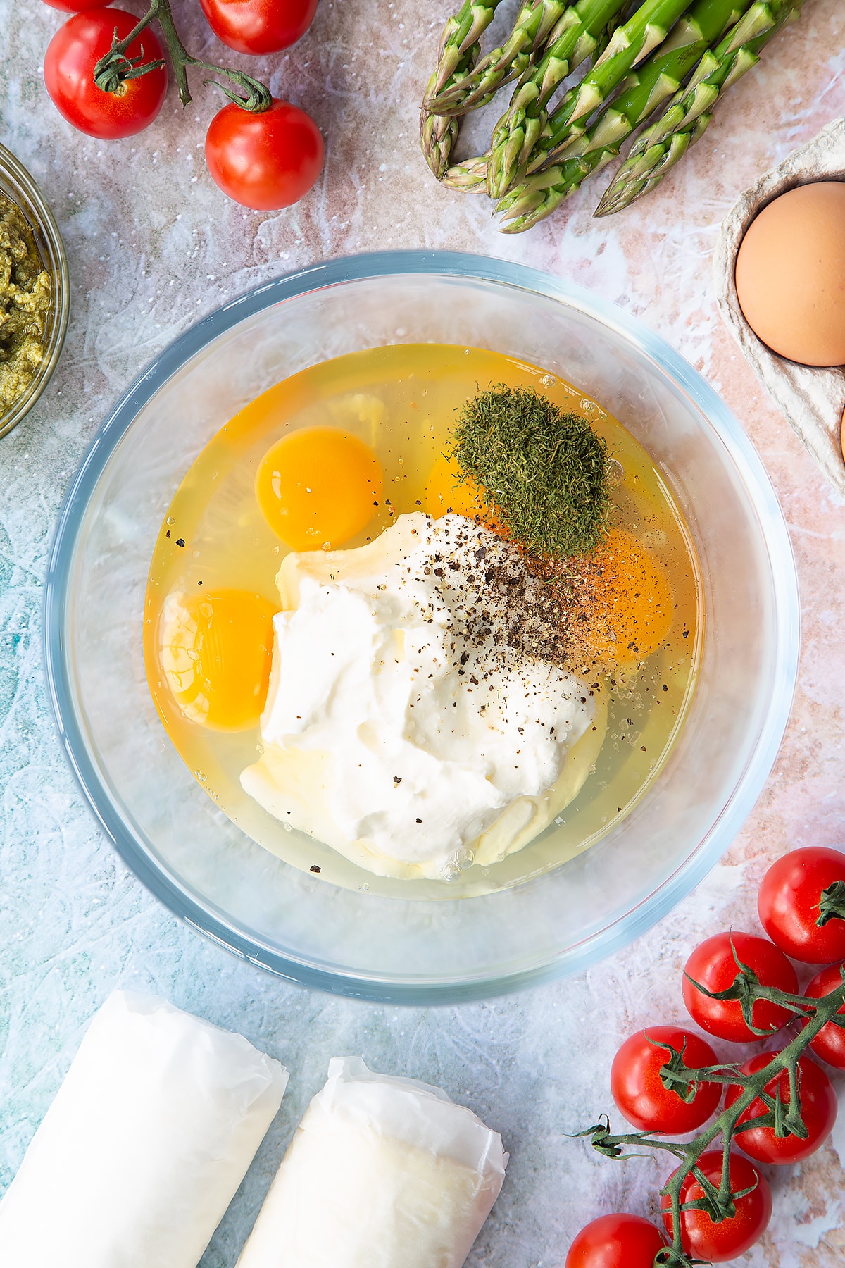 Eggs, crème fraîche, dill and a pinch of salt and pepper in a glass bowl. Ingredients to make asparagus tartlets surround the bowl.