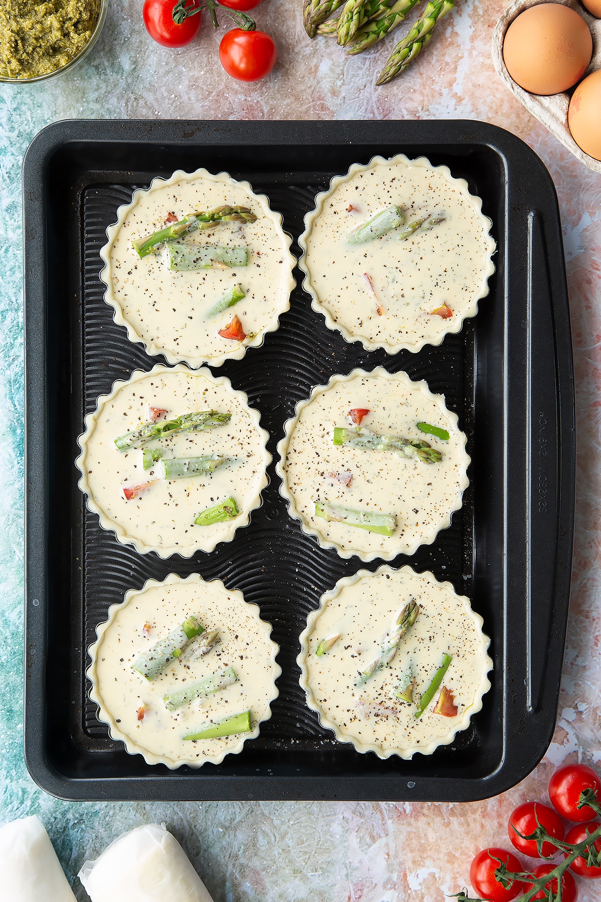 Six tartlet tins on a baking tray. Pastry is fitted in the tins, which is filled with tart filling, cherry tomato and asparagus pieces. Ingredients to make asparagus tartlets surround the tray.