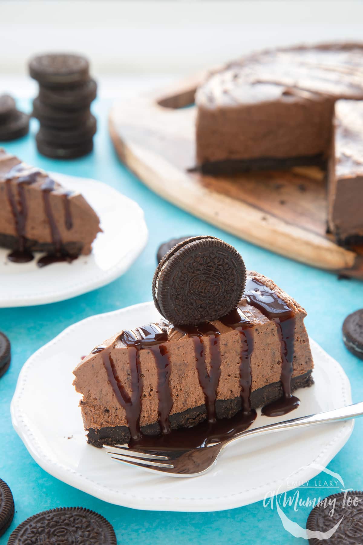 Front view shot of a piece of chocolate oreo cheesecake on a white plate drizzled with melted chocolate sauce topped with a oreo cookie