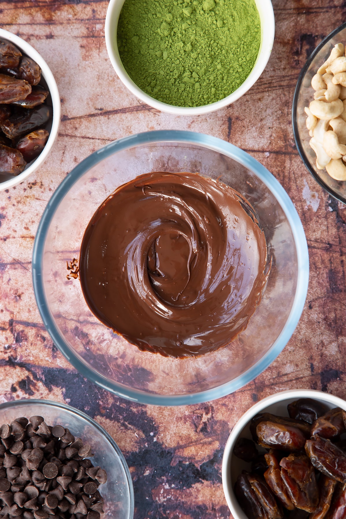 Melted dark chocolate in a mixing bowl. Ingredients to make matcha protein balls surround the bowl.
