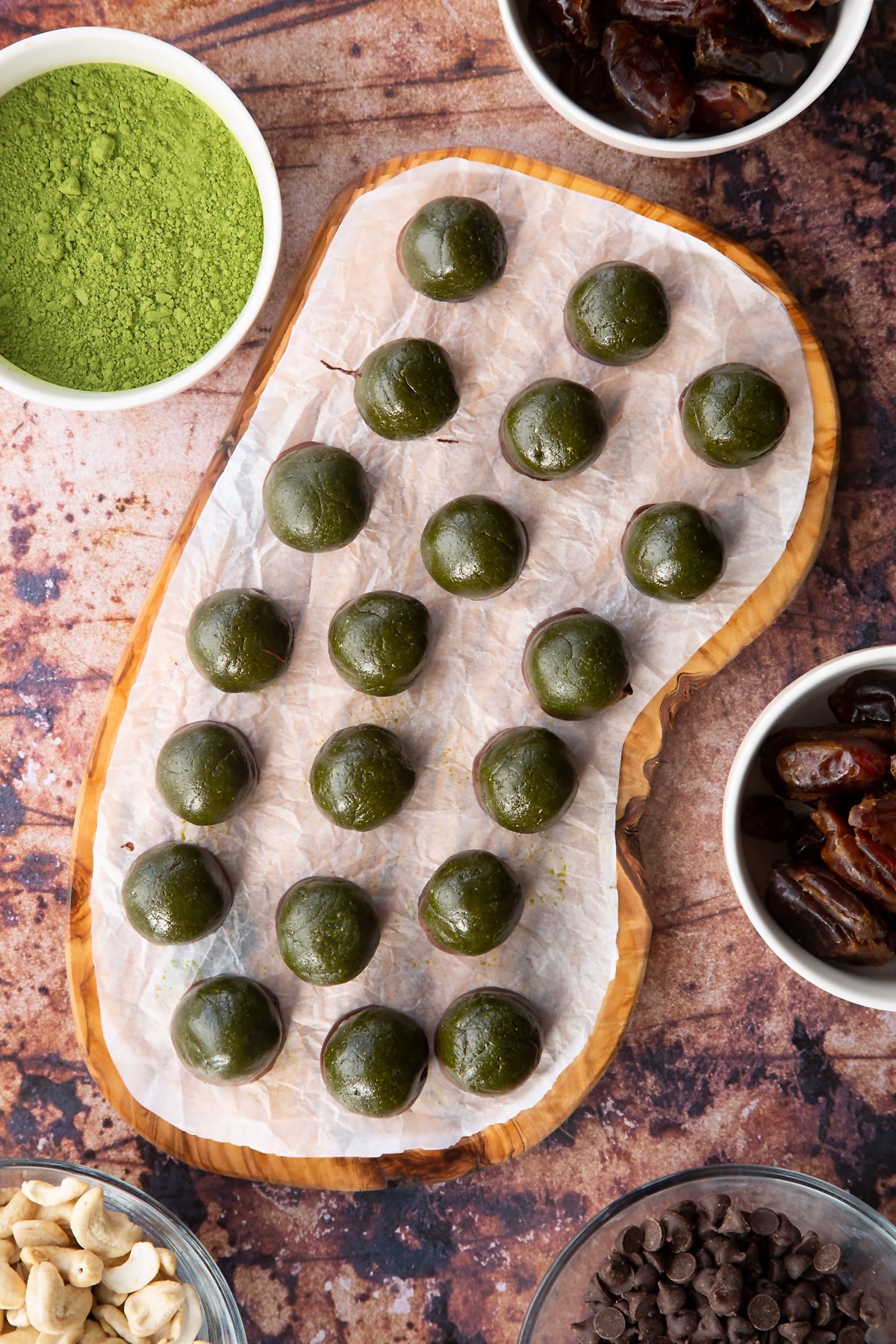 Matcha protein balls shown from above on a wooden board lined with baking paper. The balls are dark green and the bases have been dipped in dark chocolate.