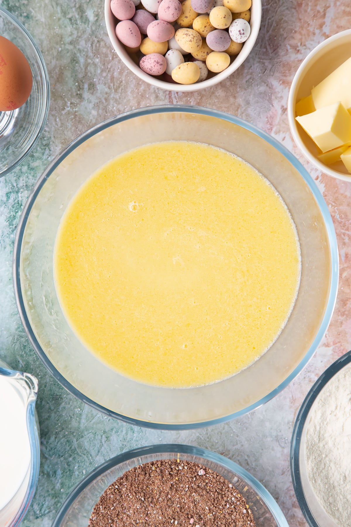 Melted butter, milk and eggs whisked together in a bowl. Ingredients to make Mini egg waffles surround the bowl.