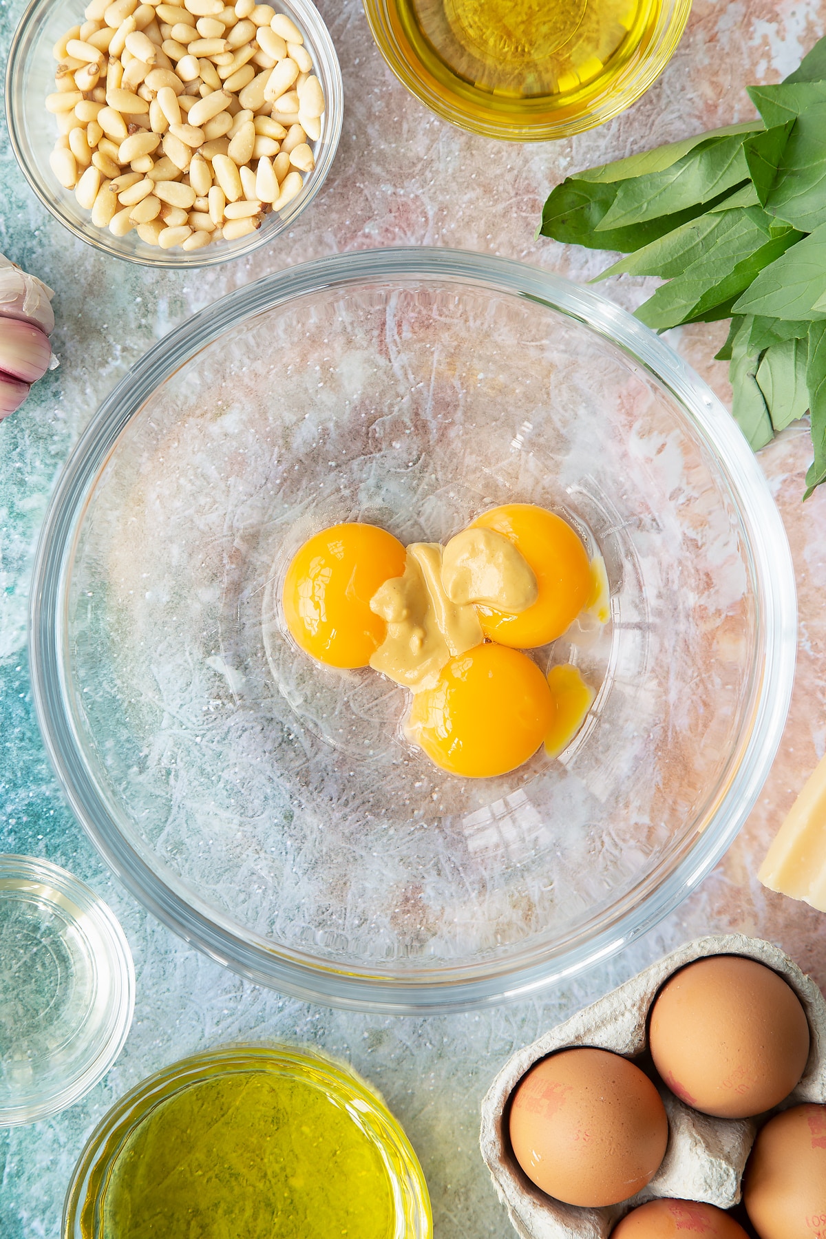 Egg yolks, mustard and vinegar in a bowl. Ingredients to make pesto mayo surround the bowl.