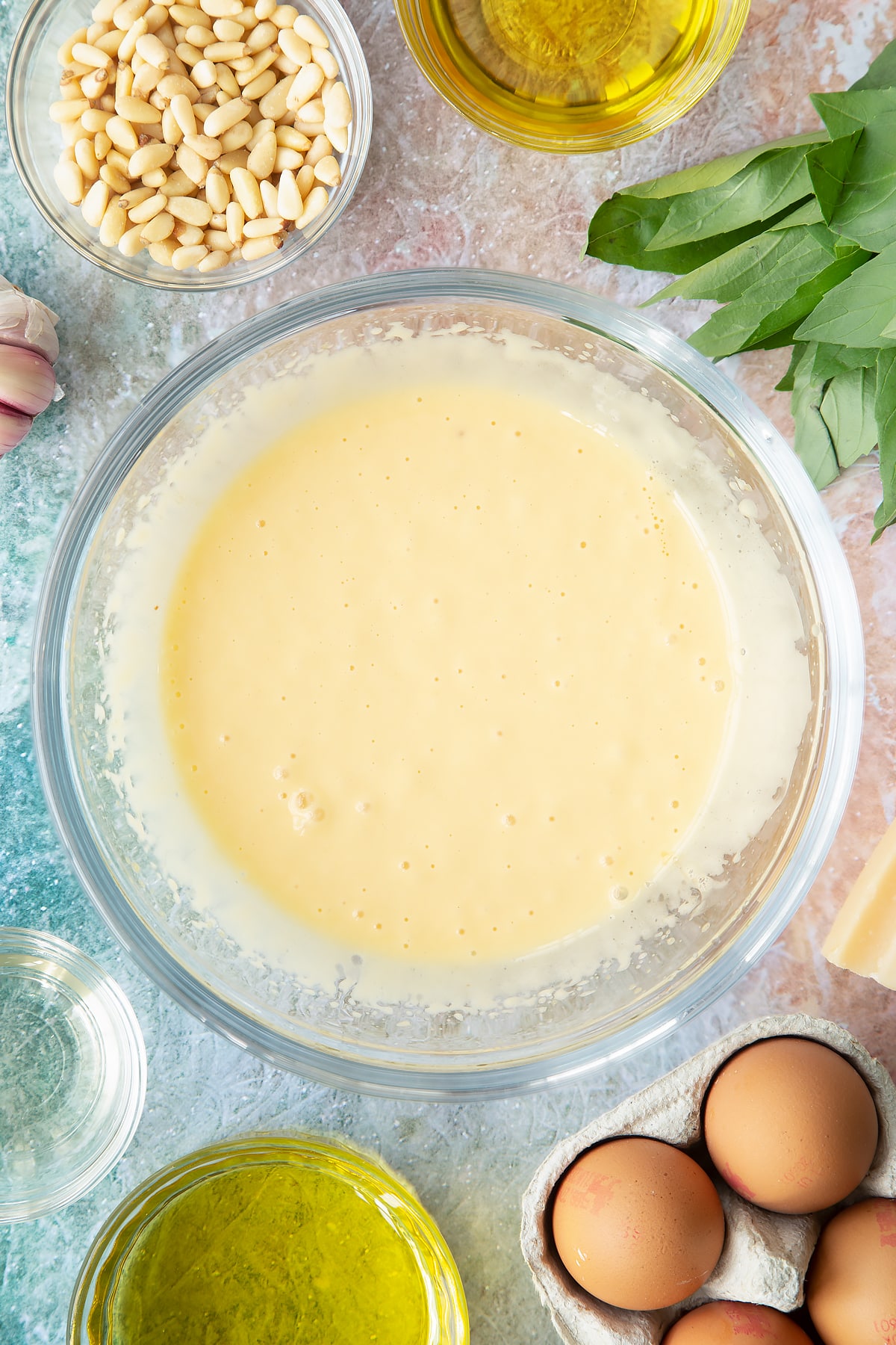 Egg yolks, mustard, vinegar and a little oil whisked together in a bowl. Ingredients to make pesto mayo surround the bowl.
