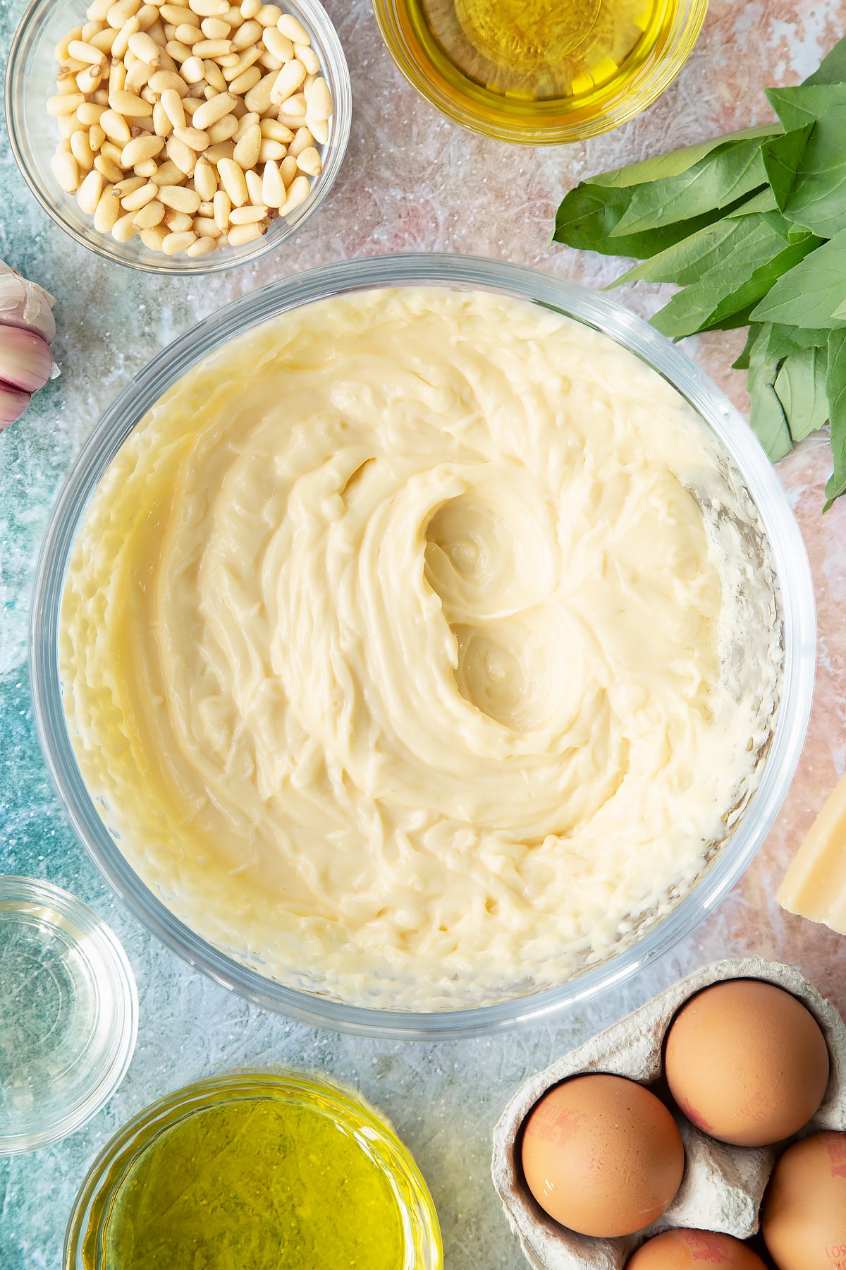 Freshly whisked mayo in a bowl. Ingredients to make pesto mayo surround the bowl.