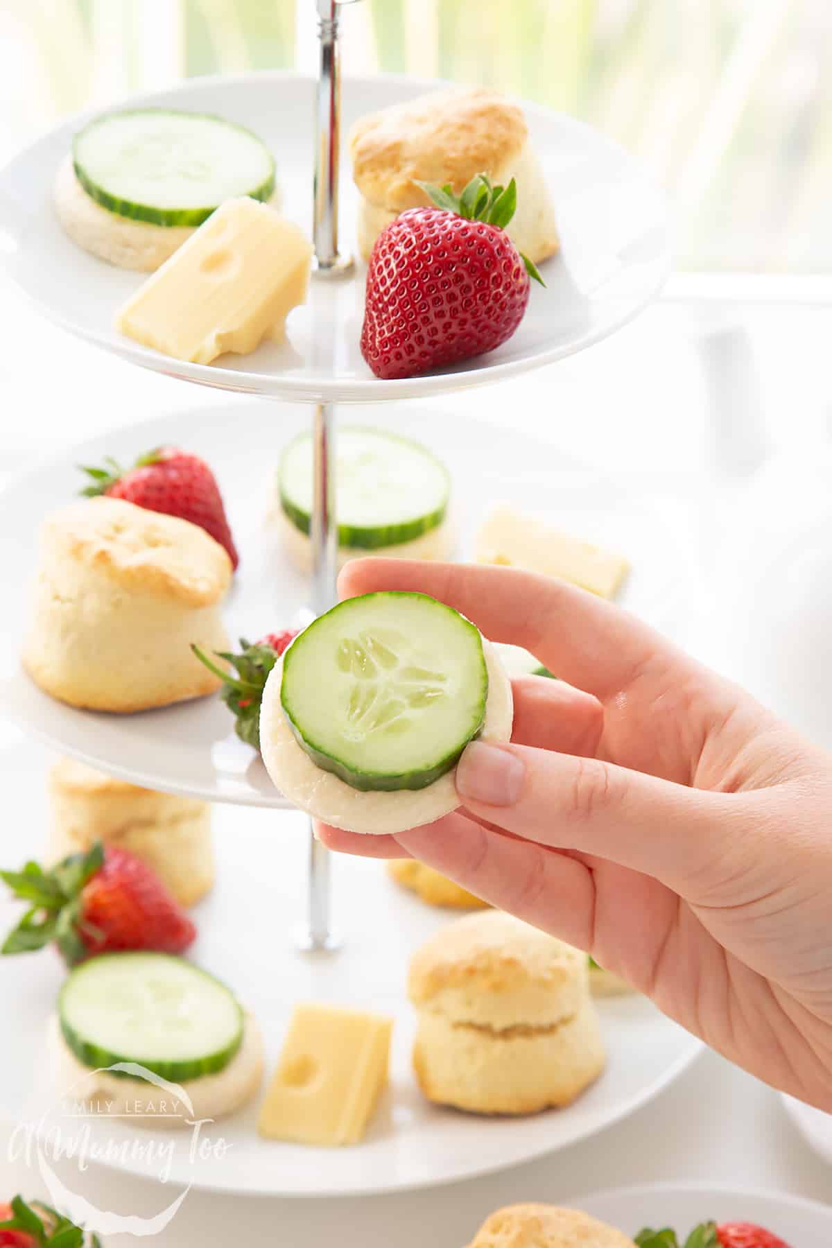 Hand holding one of the sandwhiches with the three tier cake stand filled with sandwhiches, scones and cheese in the background.