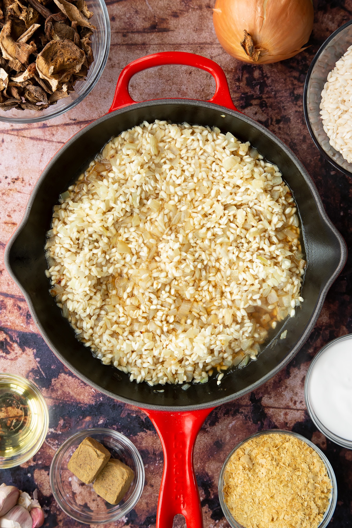 Lightly fried onions and arborio rice covered with a little stock in a pan. Ingredients to make vegan mushroom risotto surround the pan.