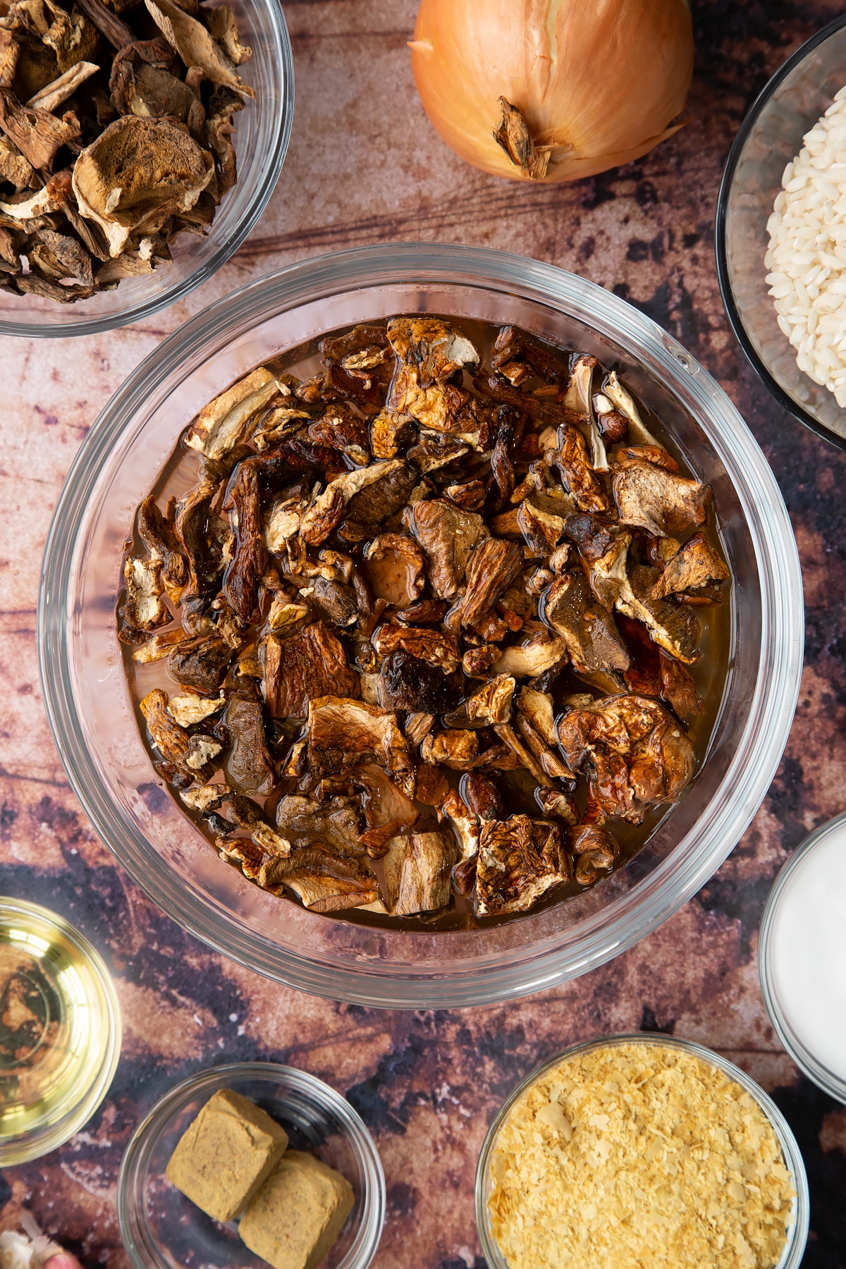 Dried porcini mushrooms covered with water in a glass bowl. Ingredients to make vegan mushroom risotto surround the bowl.