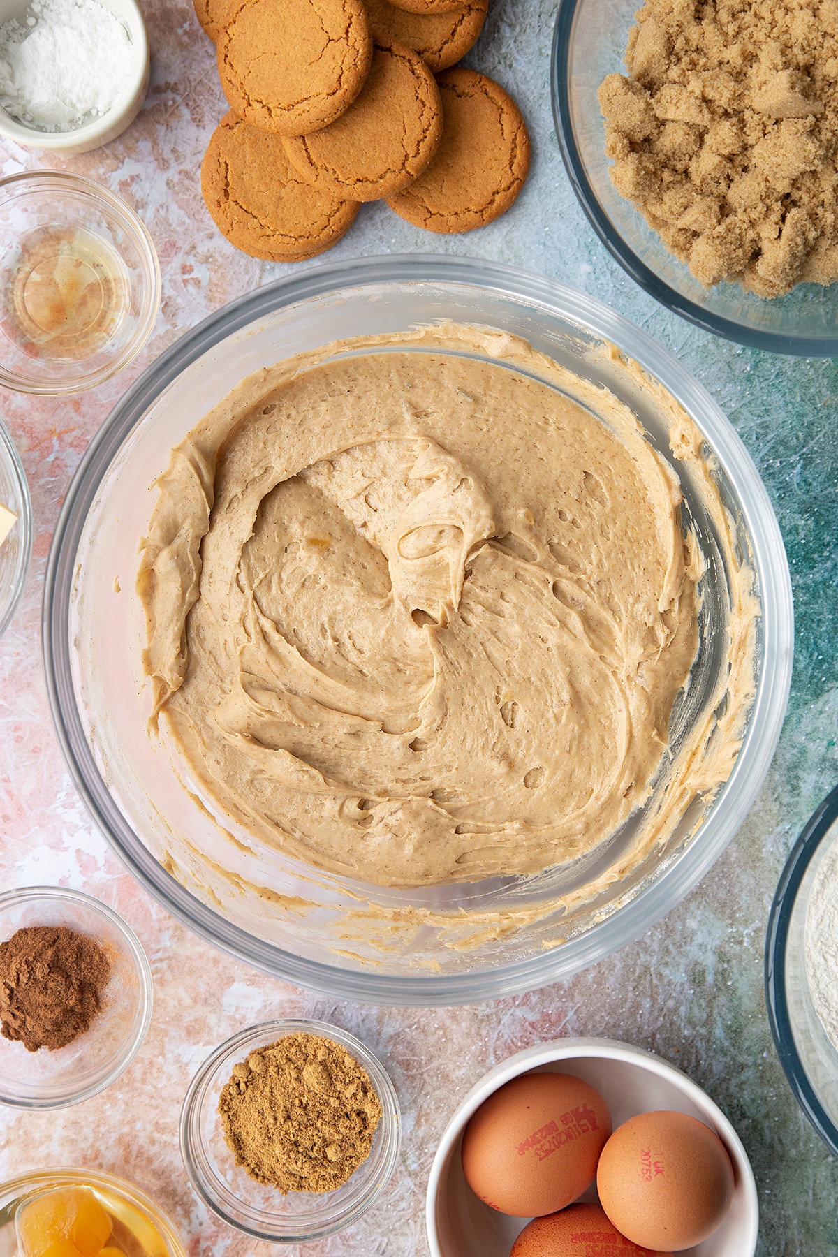 Ginger cake batter with stem ginger in a mixing bowl. Ingredients to make ginger cupcakes surround the bowl.