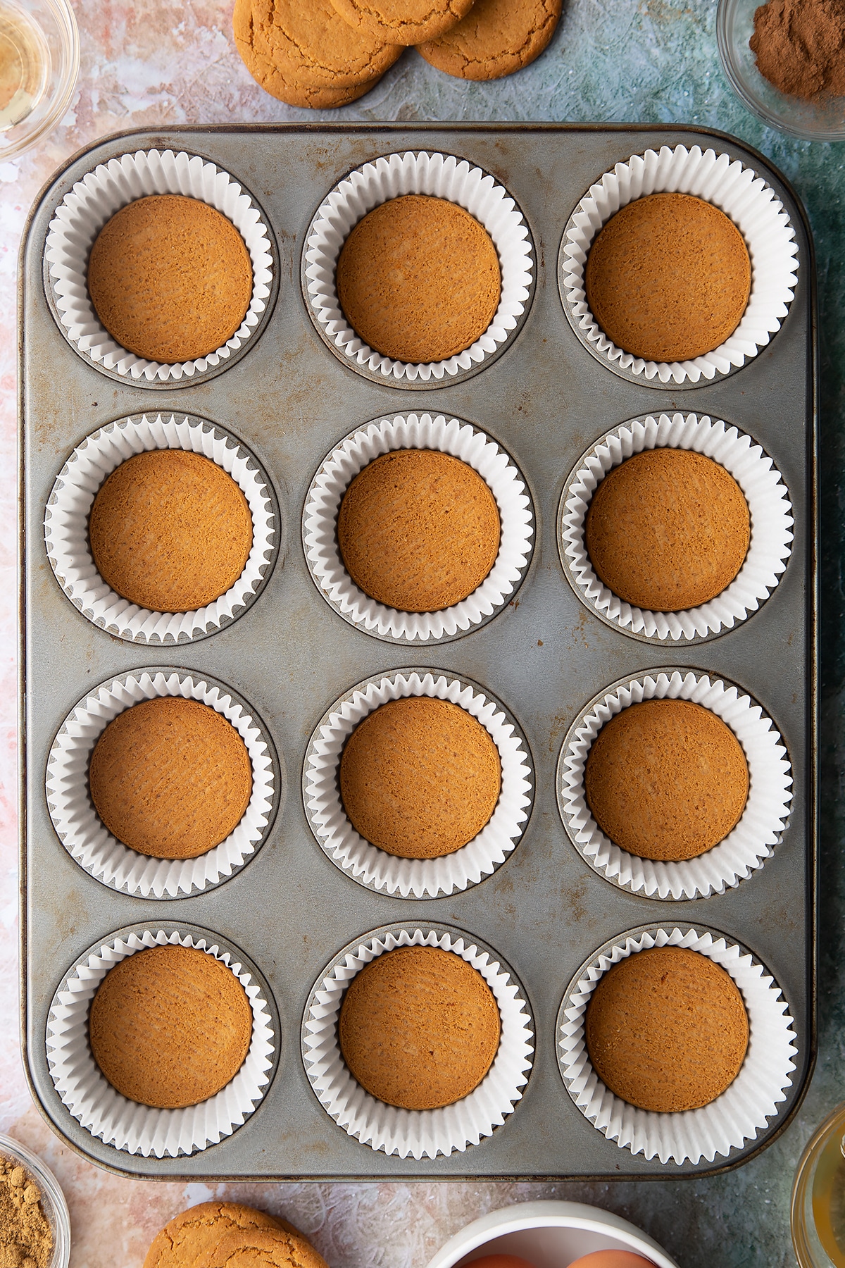 Muffin tin lined with cupcake cases. There's a gingernut biscuit in the base of each case. Ingredients to make ginger cupcakes surround the tin.