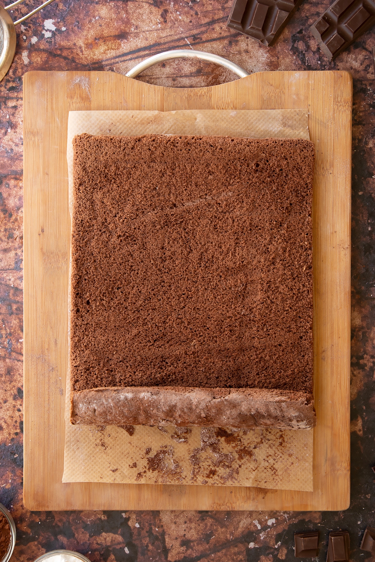 A chocolate sponge unrolled on a wooden board and baking paper dusted with cocoa and icing sugar.