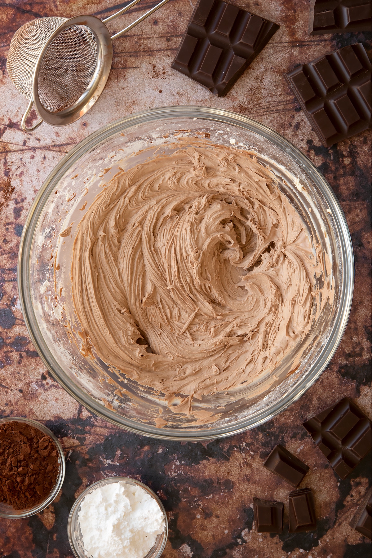Chocolate buttercream in a bowl. Ingredients to make chocolate Swiss roll surround the bowl.