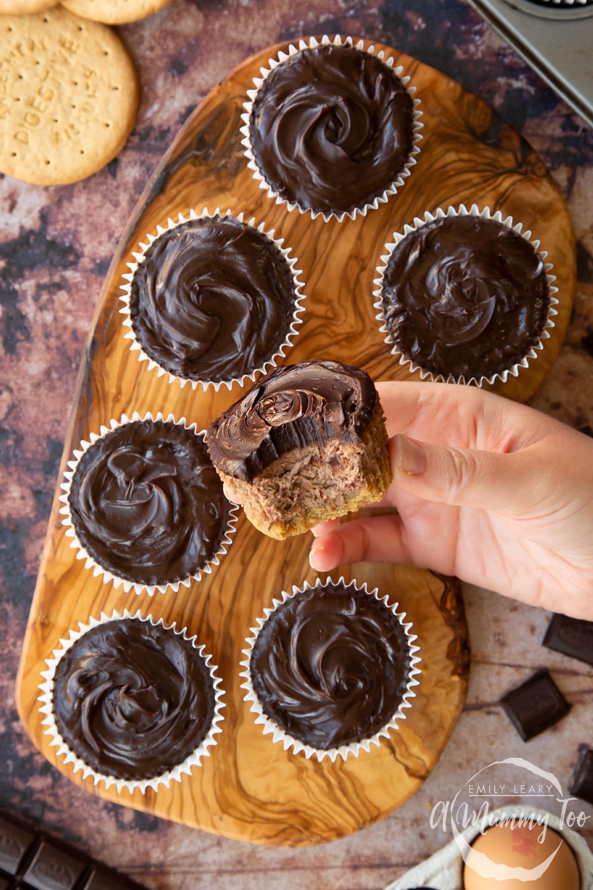 Chocolate cheesecake cupcakes in rows on an olive wood board, shown from above. A hand holds one with a bite out of it.
