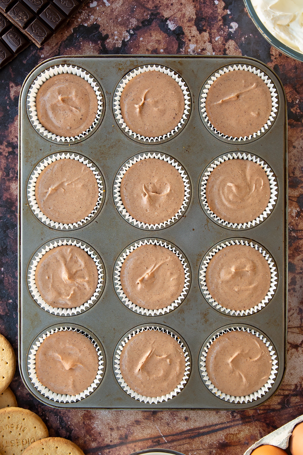 Chocolate cheesecake mix in cupcake cases in a muffin tray. Ingredients to make chocolate cheesecake cupcakes surround the tray.