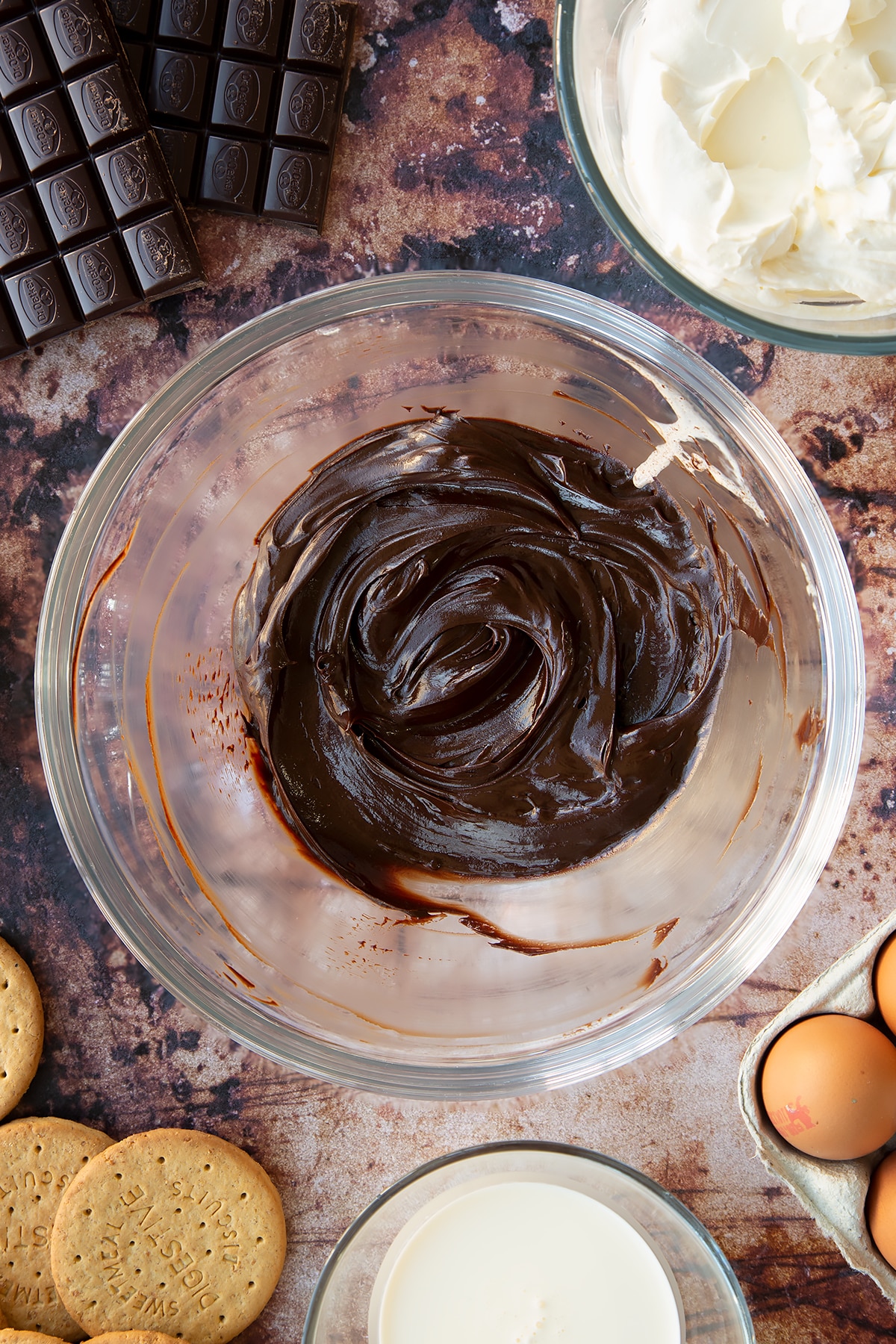 Dark chocolate ganache in a glass bowl. Ingredients to make chocolate cheesecake cupcakes surround the bowl.