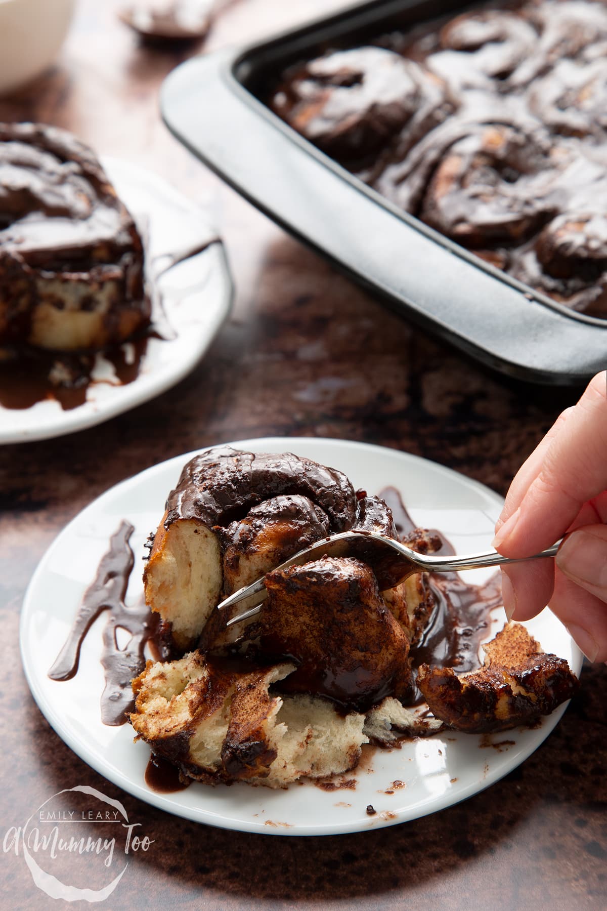 Front view of a hand holding a fork cutting into a chocolate cinnamon bun on a white plate with the A Mummy Too Logo in the corner