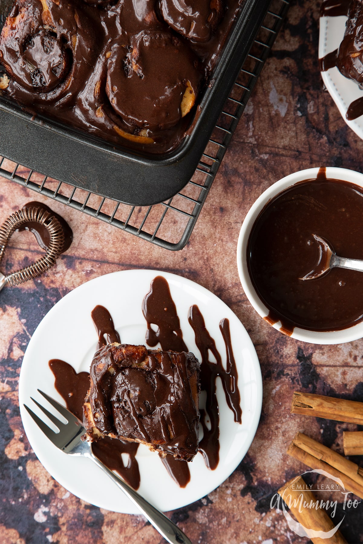 Overhead shot of a chocolate cinnamon bun drizzled with melted chocolate on a white plate with a fork with the A Mummy Too logo in the corner