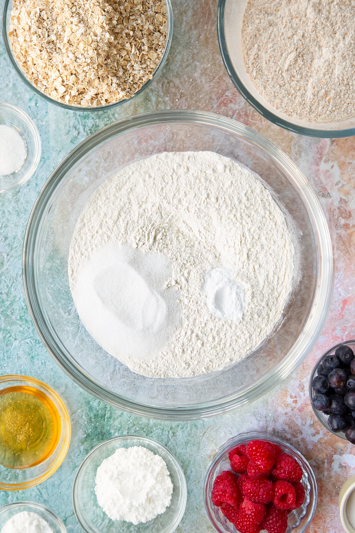 Self-raising flour, bicarbonate of soda and sugar in a glass mixing bowl. Ingredients to make oatmeal scones surround the bowl.