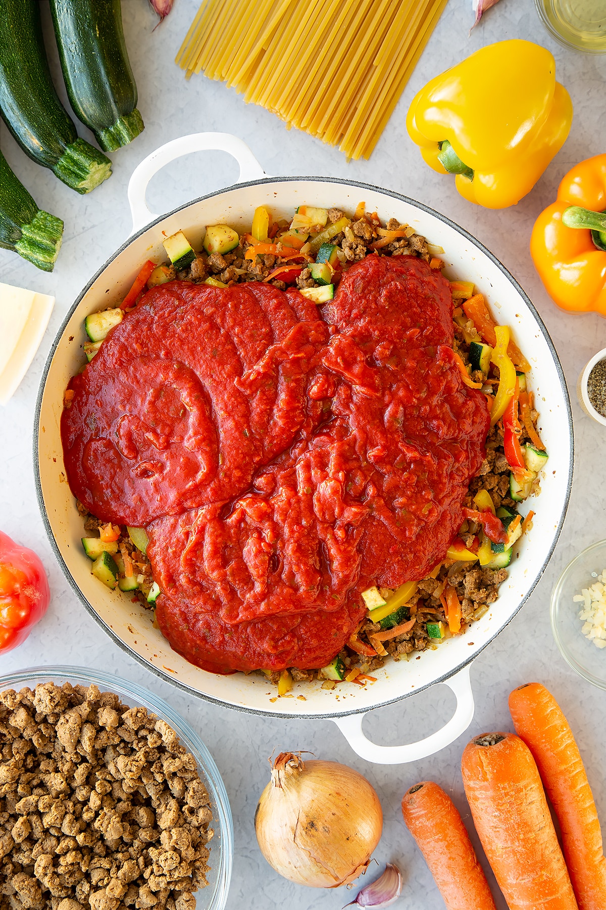 Veggie mince, garlic, onion, courgette, carrot and peppers sweated in a large pan with pasta sauce on top. Ingredients to make one pot vegan bolognese surround the pan.