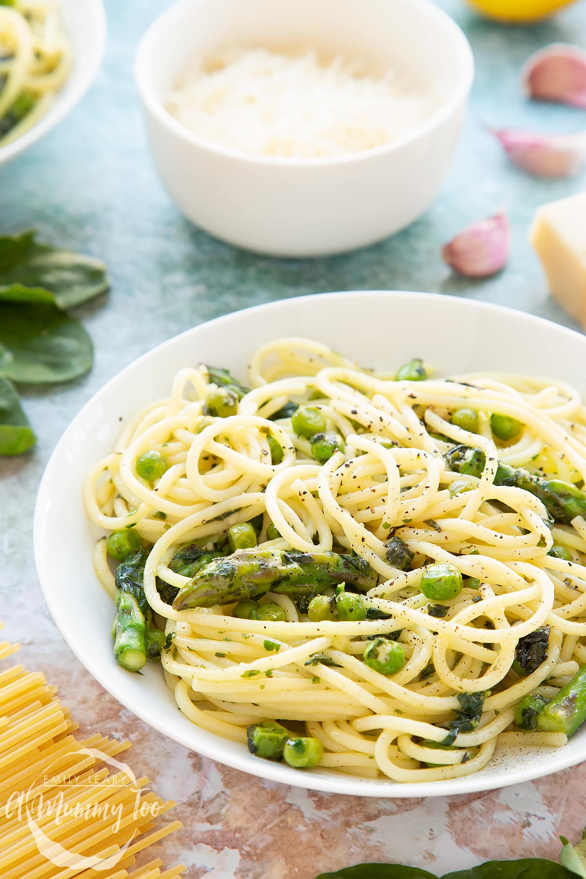 Summer spaghetti with spinach, basil, asparagus and peas, served in a white bowl. A bowl of parmesan sits in the background.