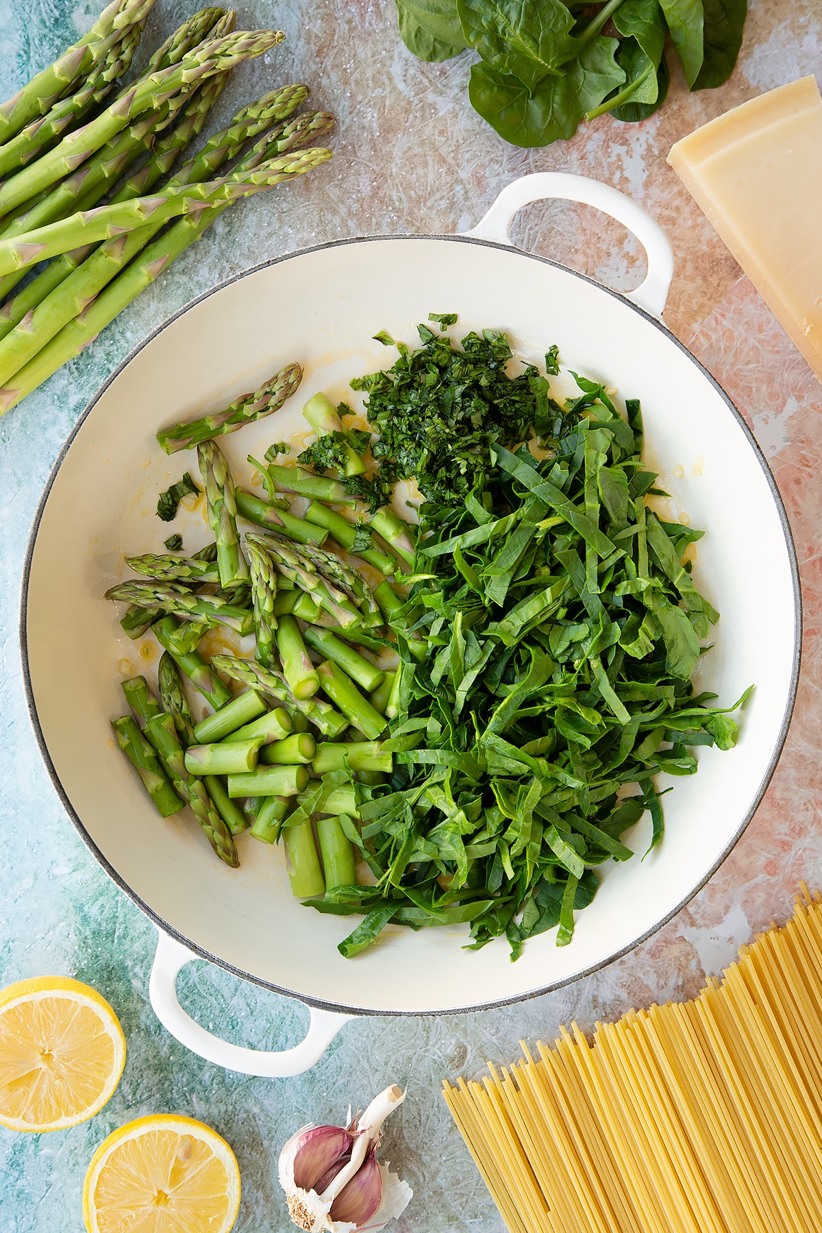 Fresh spinach, basil and asparagus in a large, shallow saucepan. Ingredients to make summer spaghetti surround the pan.
