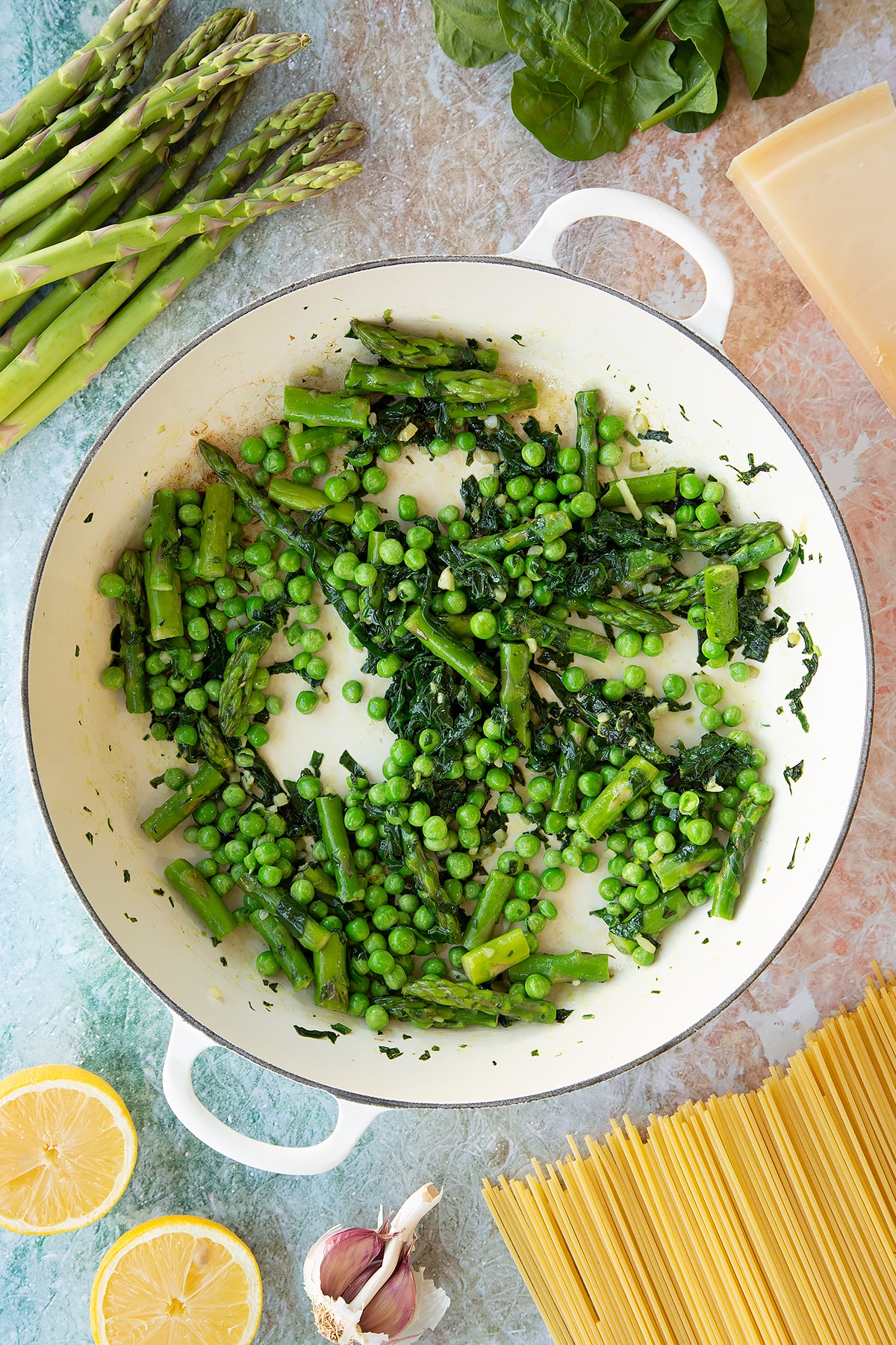Lightly sautéed spinach, basil, asparagus and peas in a large, shallow saucepan. Ingredients to make summer spaghetti surround the pan.