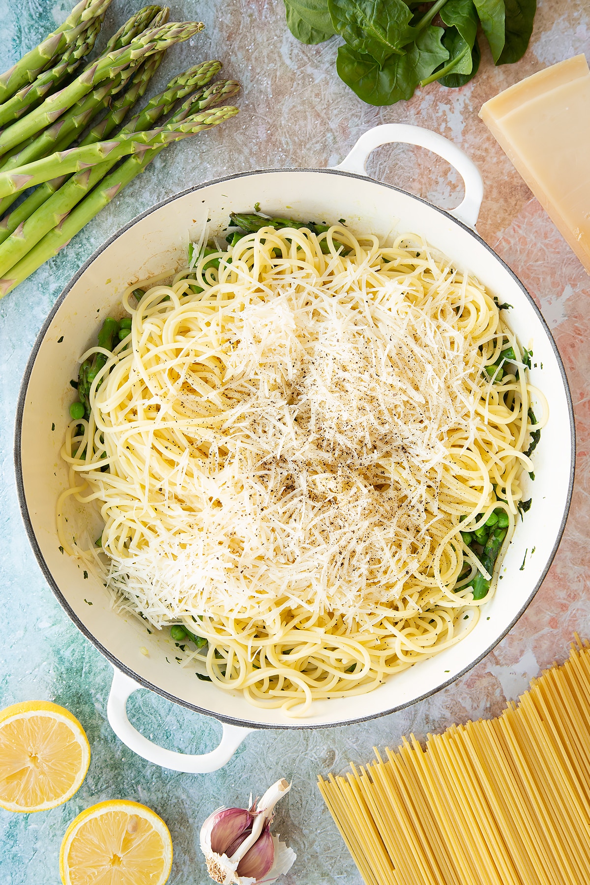 Lightly sautéed spinach, basil, asparagus and peas in a large, shallow saucepan with spaghetti and parmesan on top. Ingredients to make summer spaghetti surround the pan.