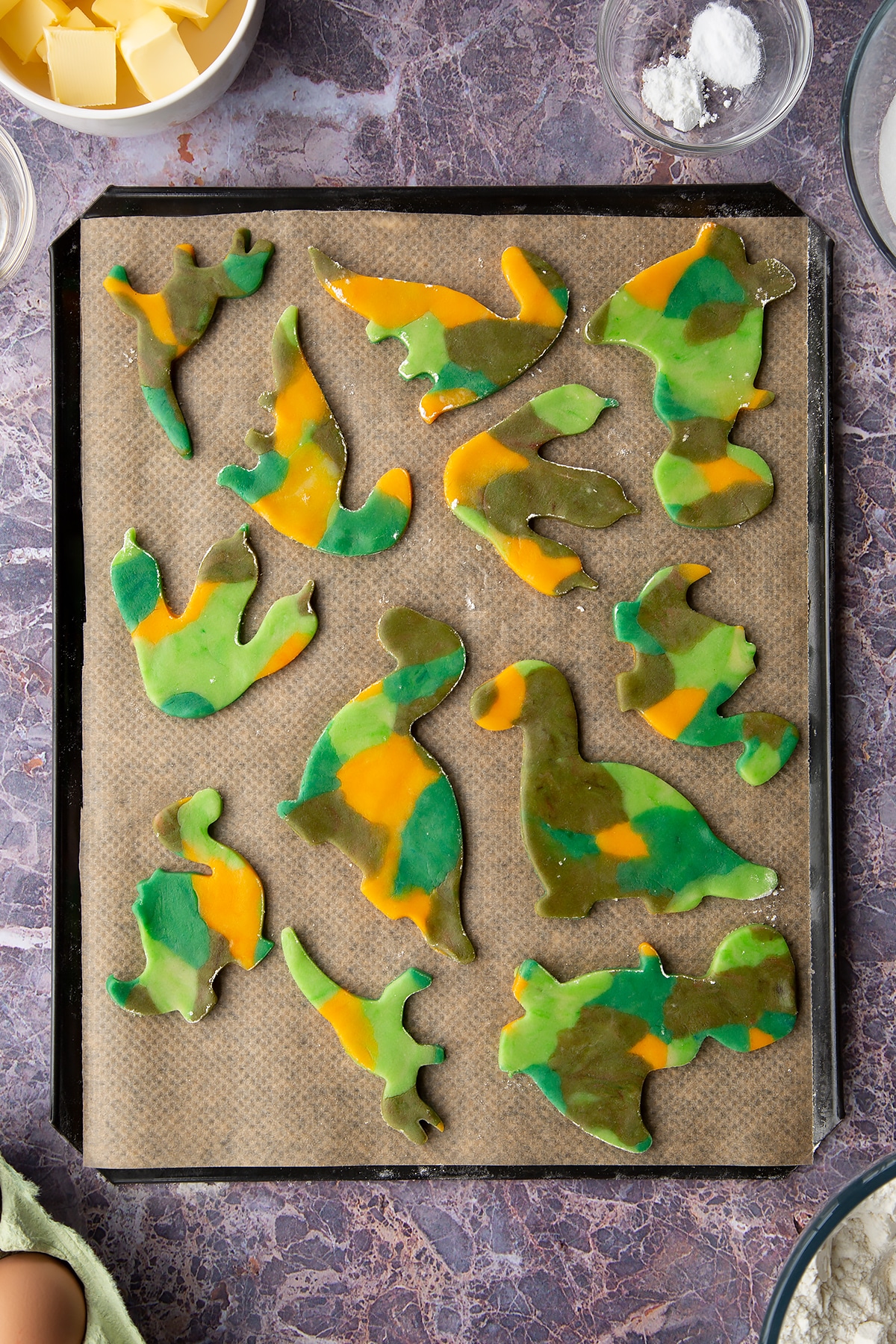Overhead shot of multicoloured dinosaur cookie dough shapes on a lined baking tray