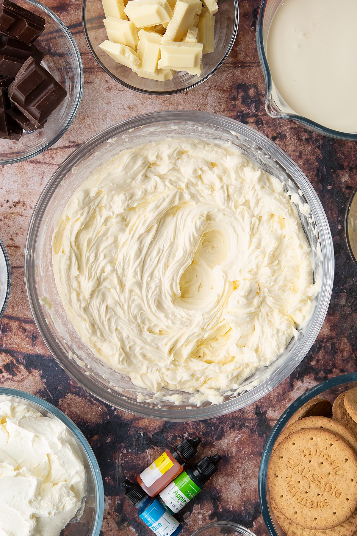 Double cream, cream cheese, peppermint extract and sugar, whisked together in a glass bowl. Ingredients to make no bake mint cheesecake surround the bowl.