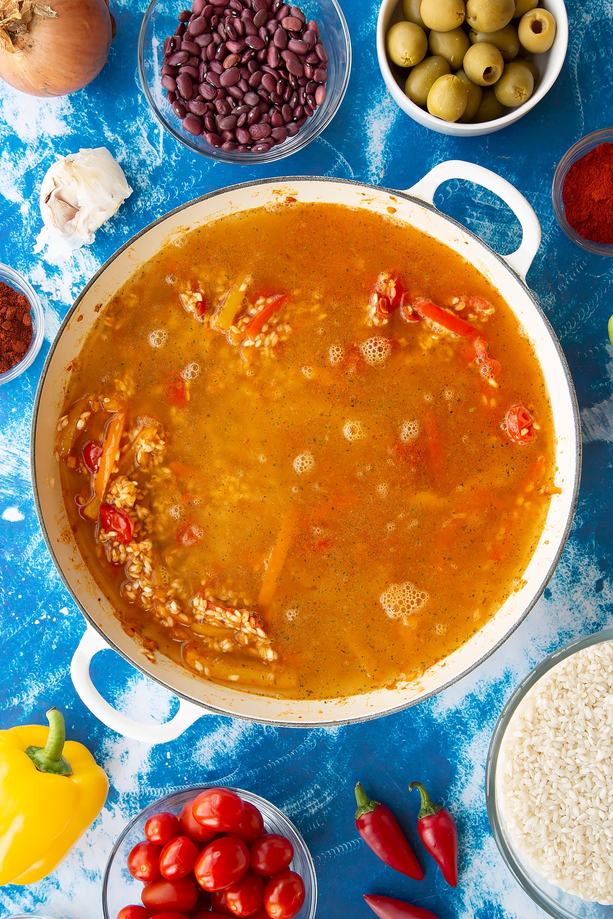 Sweated onions, spices, tomatoes and mixed sliced peppers in a pan with arborio rice and stock. Ingredients to make easy Spanish rice and beans surround the pan.