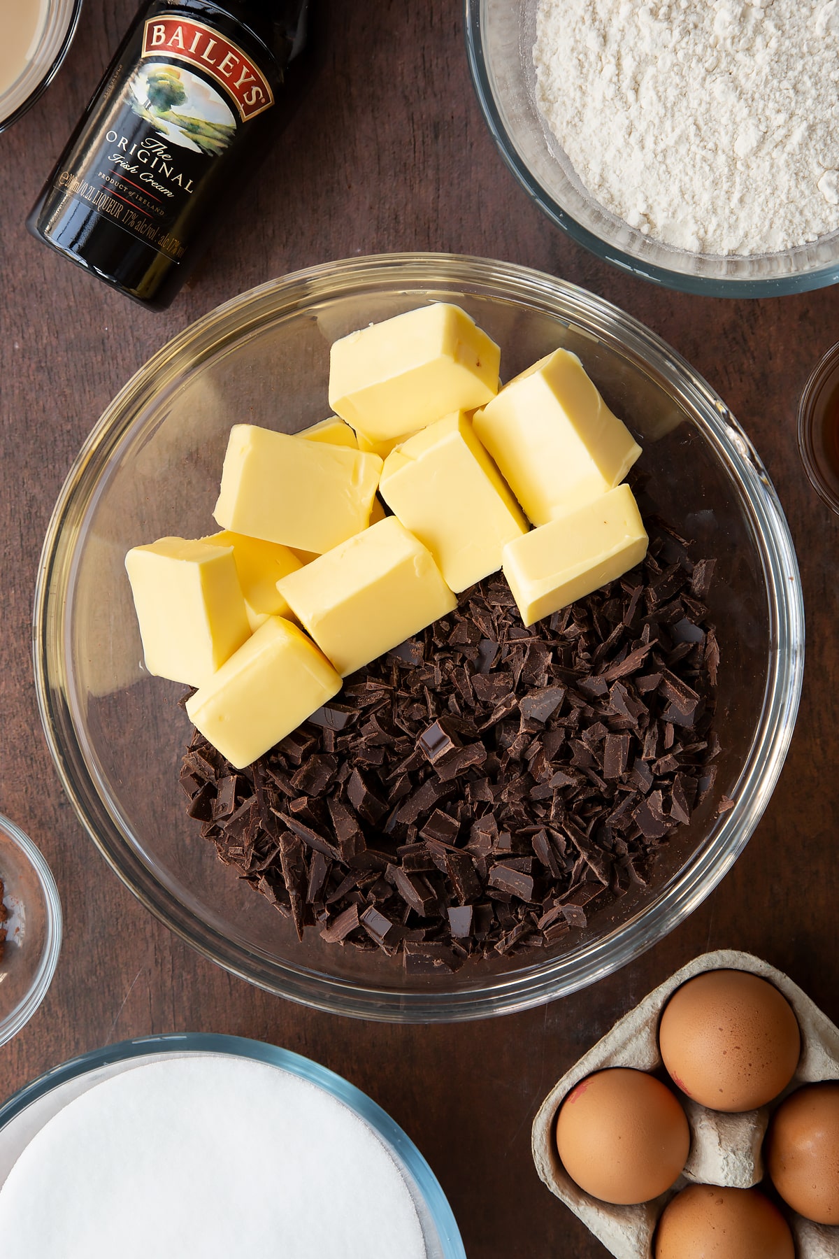 Chopped dark chocolate and cubed butter in a glass bowl. Ingredients to make Baileys brownies surround the bowl.