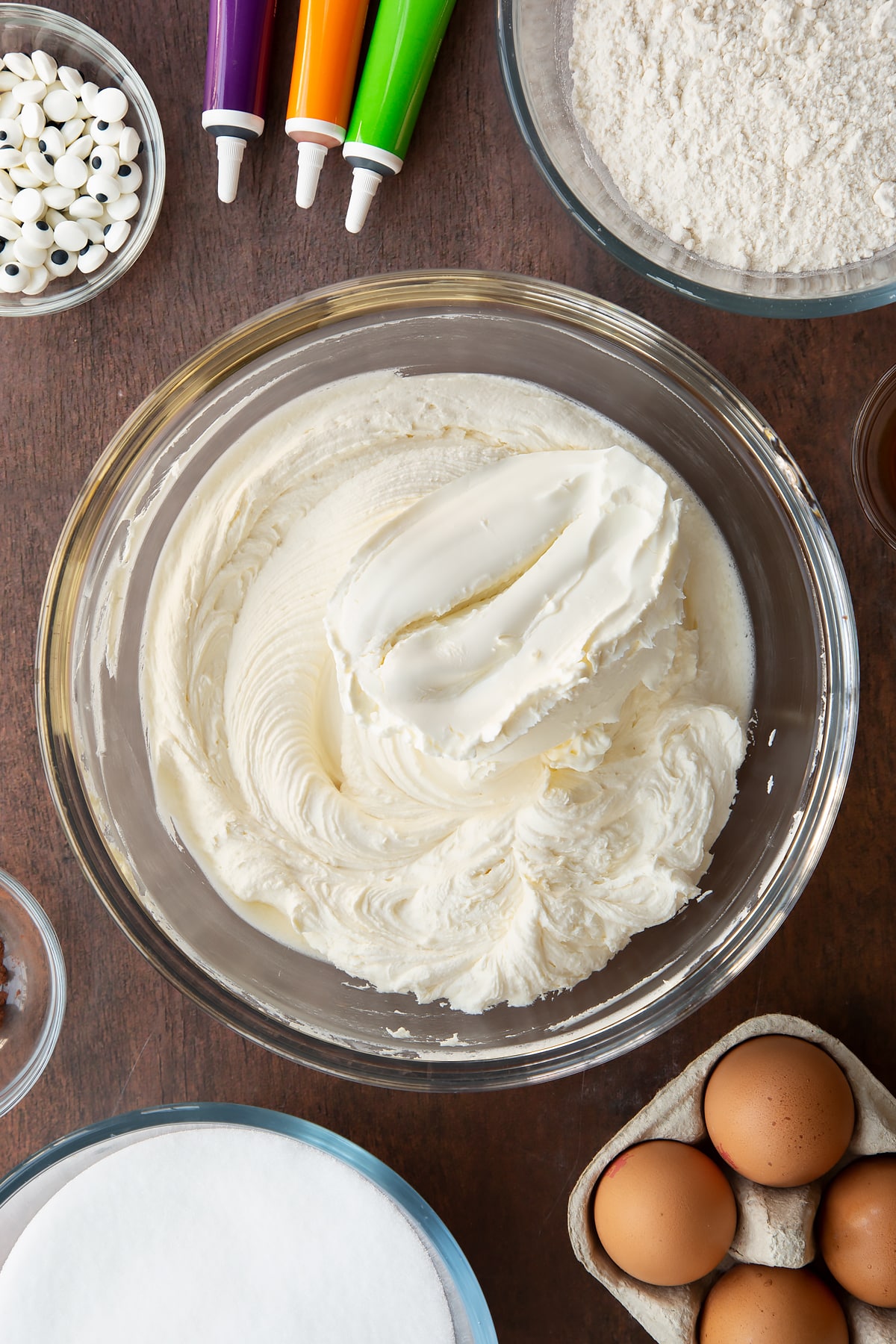 Frosting in a glass bowl with cream cheese on top. Ingredients to make Halloween brownies surround the bowl.