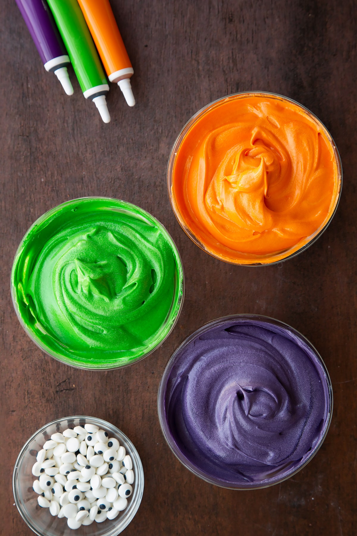 Cream cheese frosting in three glass bowls, coloured orange, green or purple.
