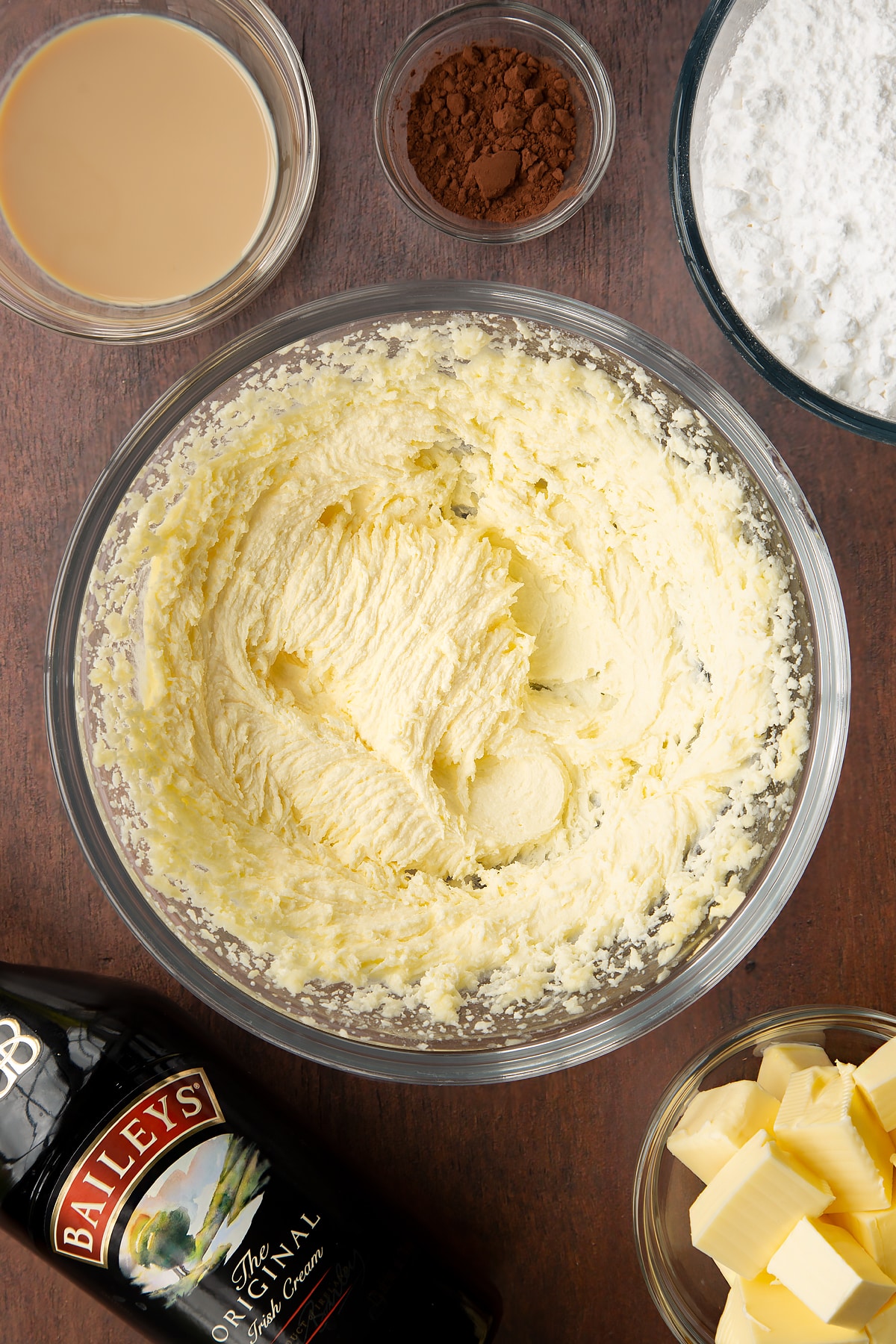 Butter, oil and sugar, whisked together in a glass mixing bowl. Ingredients to make Baileys chocolate cupcakes surround the bowl.