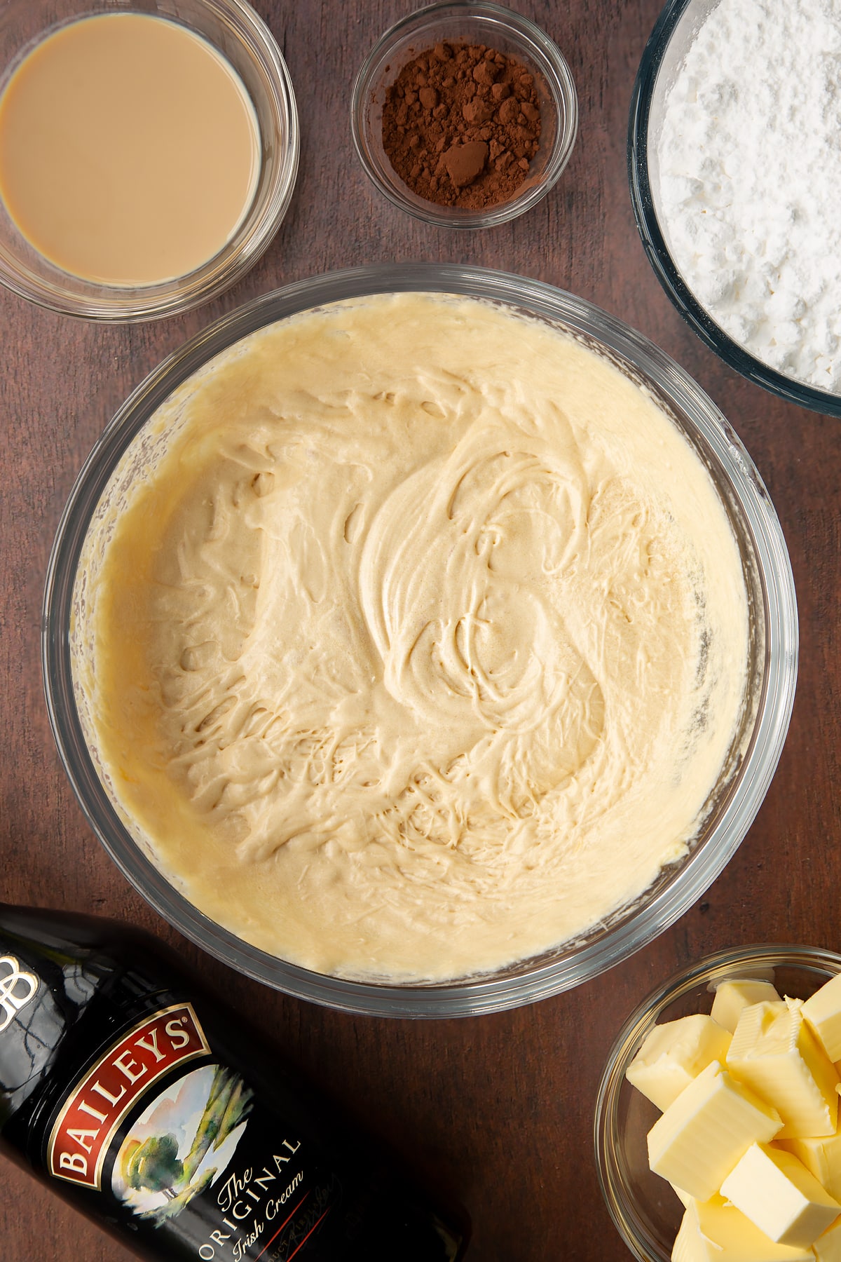 Butter, oil, sugar, eggs and Baileys whisked together in a glass mixing bowl with flour and cocoa. Ingredients to make Baileys chocolate cupcakes surround the bowl.