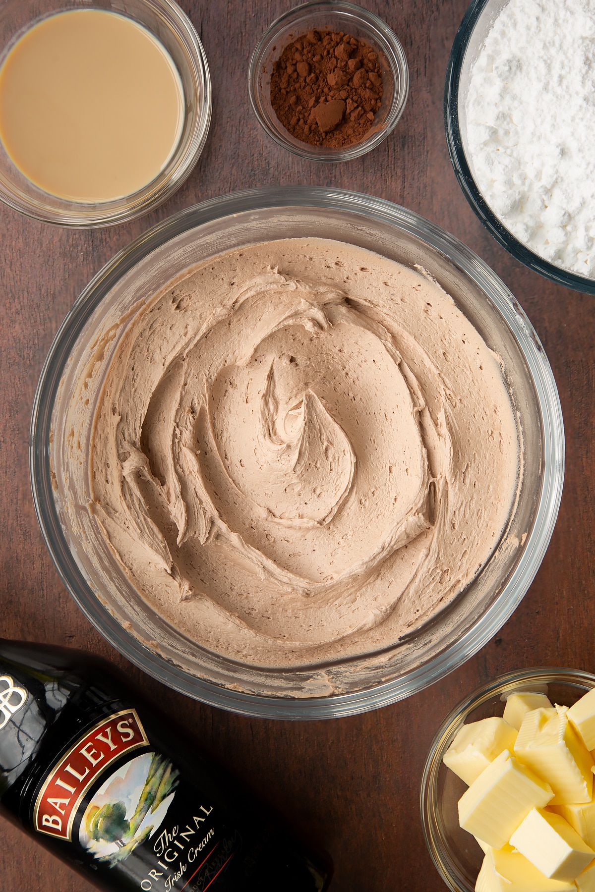 Baileys icing in a glass mixing bowl. Ingredients to make Baileys chocolate cupcakes surround the bowl.