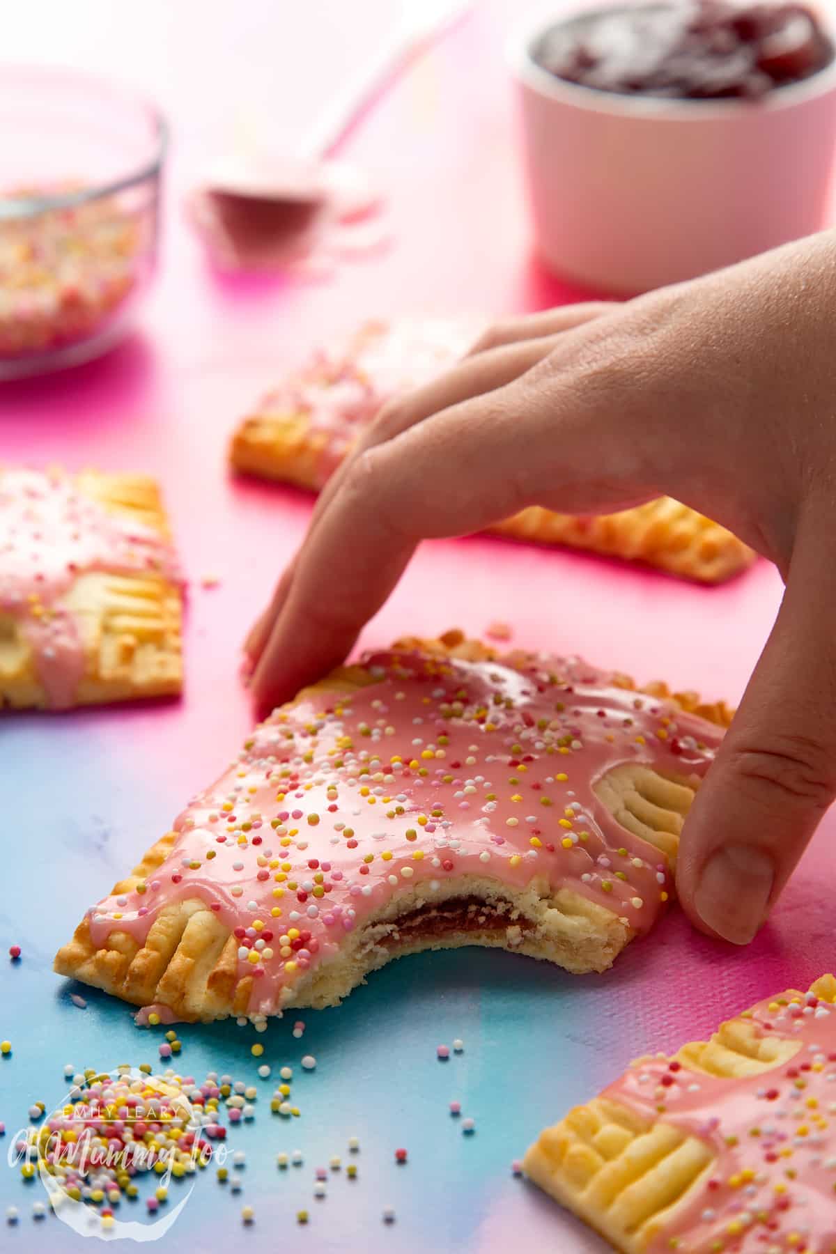 A gluten free pop tart with pink icing and pastel sprinkles. It has a bite out of it. A hand reaches for one.