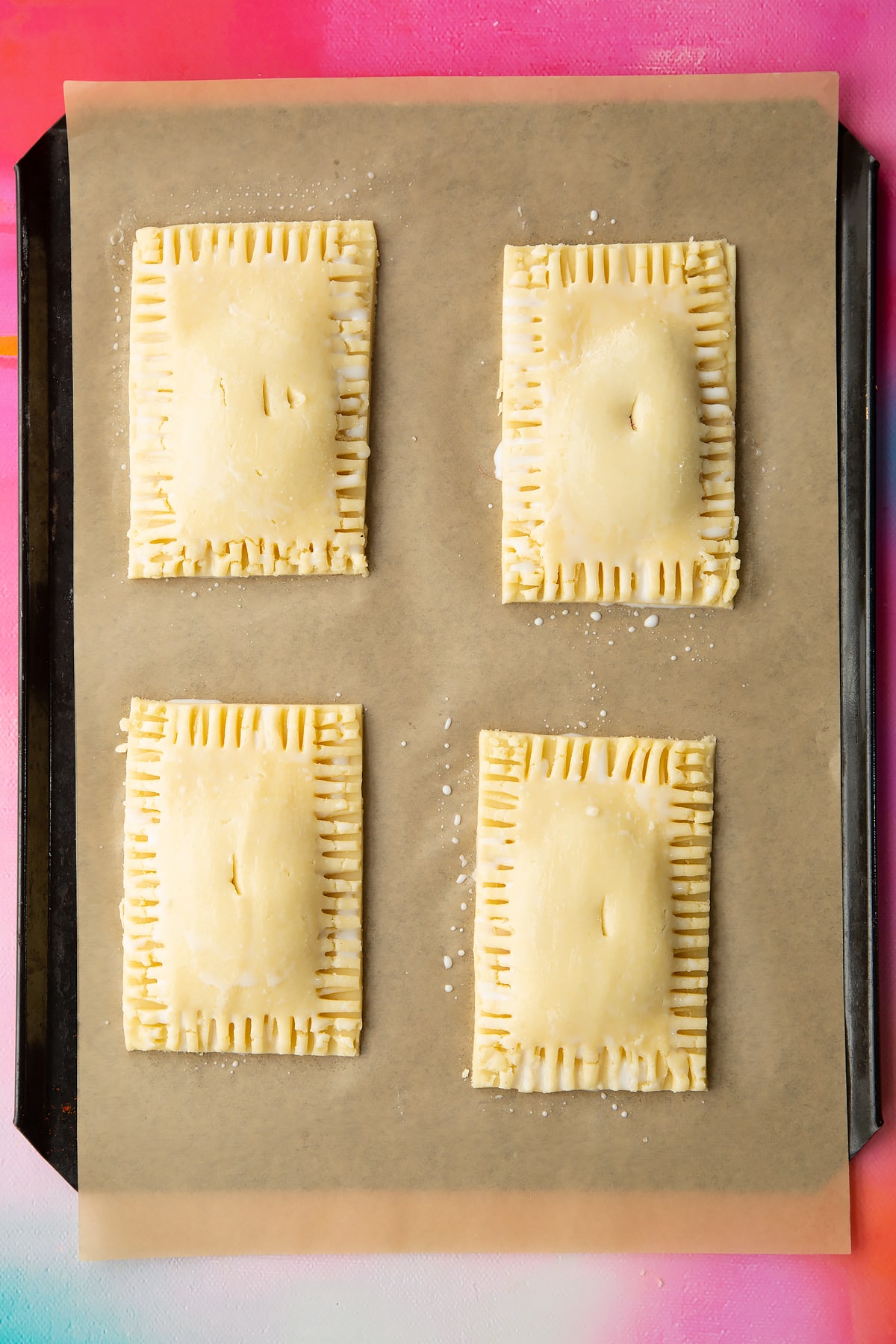 Four raw gluten free pop tarts, brushed with milk on baking tray lined with baking paper. A small hole has been cut into the top of each tart.