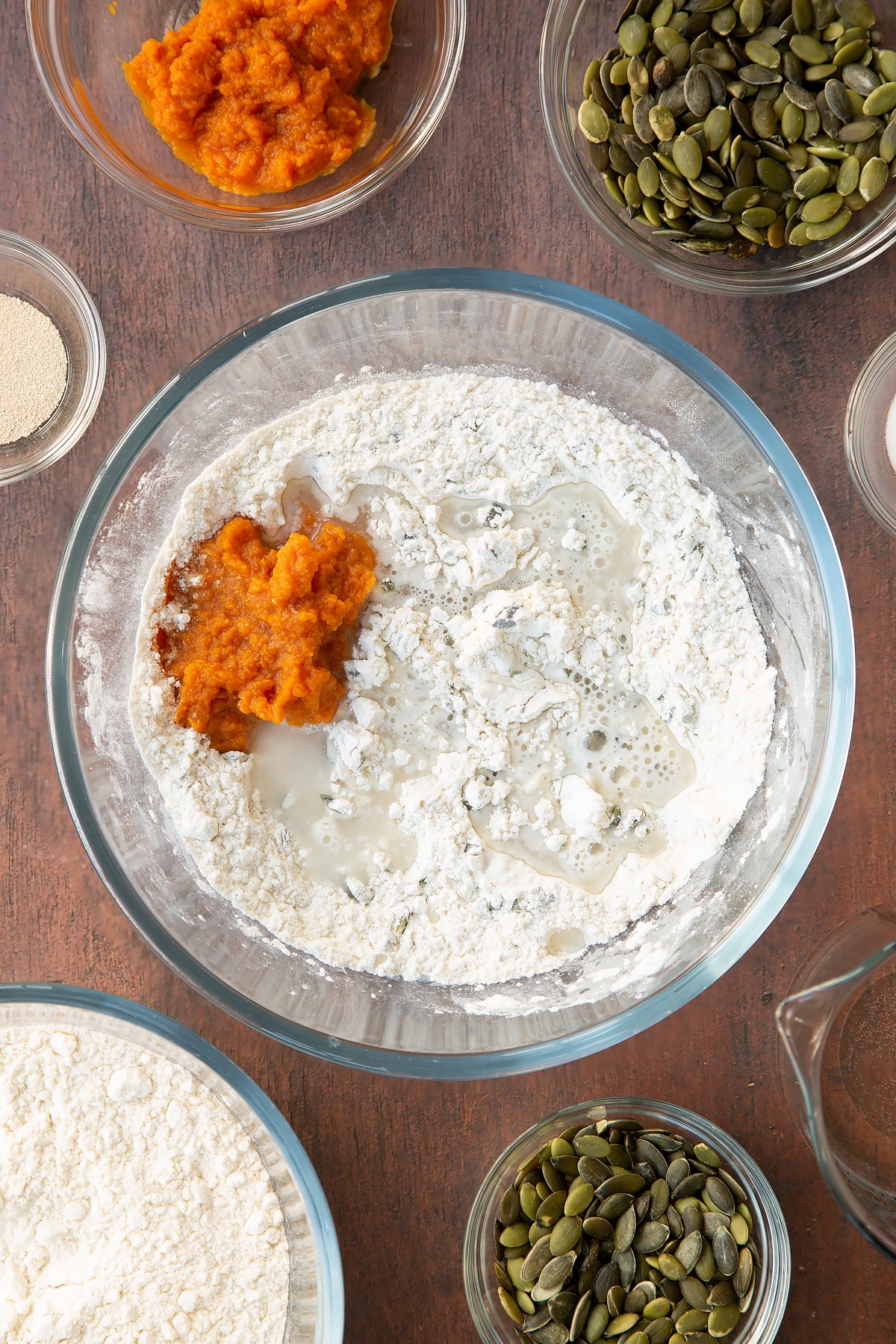 Flour, salt, yeast and pumpkin seeds mixed together in a glass mixing bowl with pumpkin puree and water on top. Ingredients to make a pumpkin seed bread recipe surround the bowl.